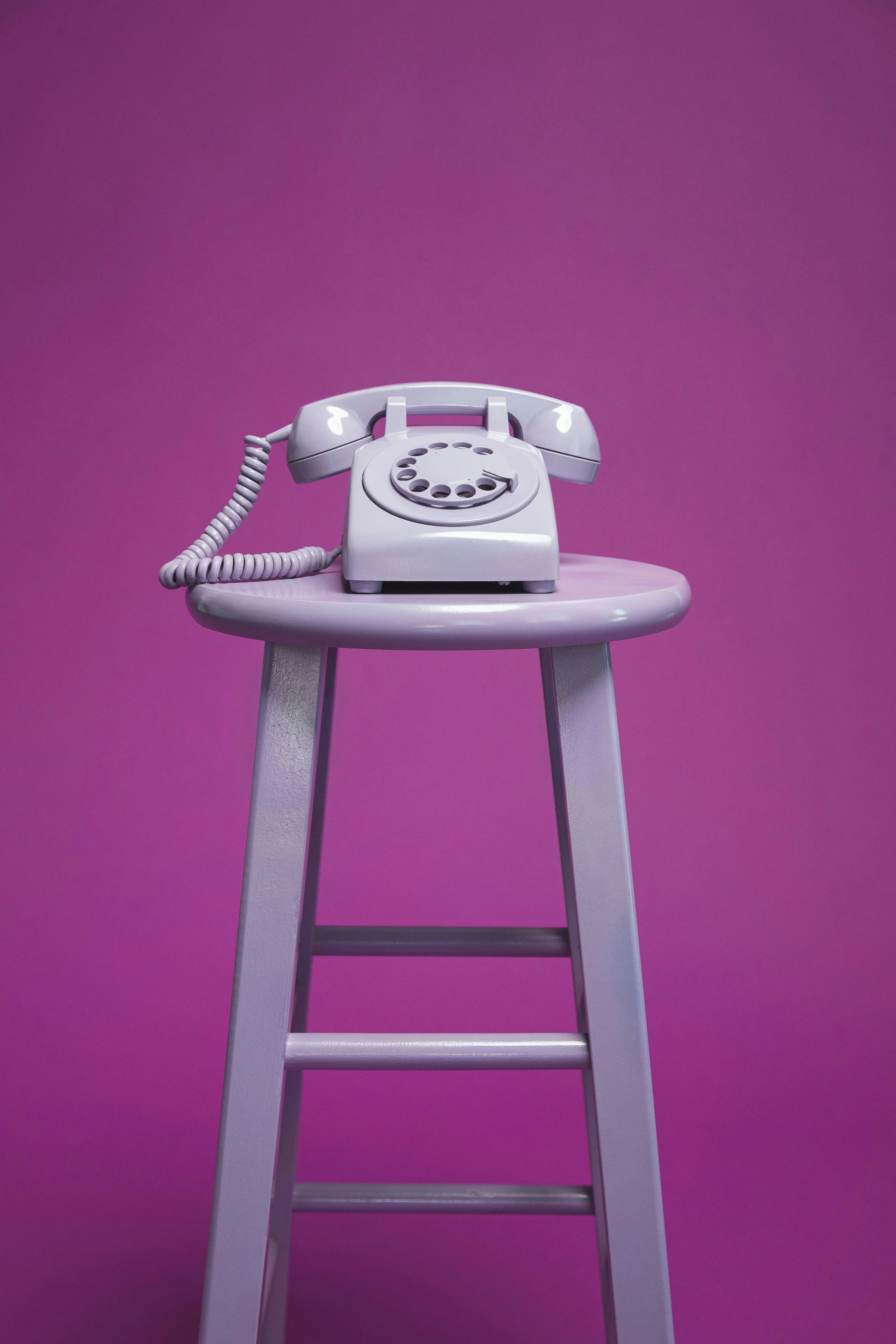 A vintage rotary telephone in purple color placed on a purple stool against a purple background.