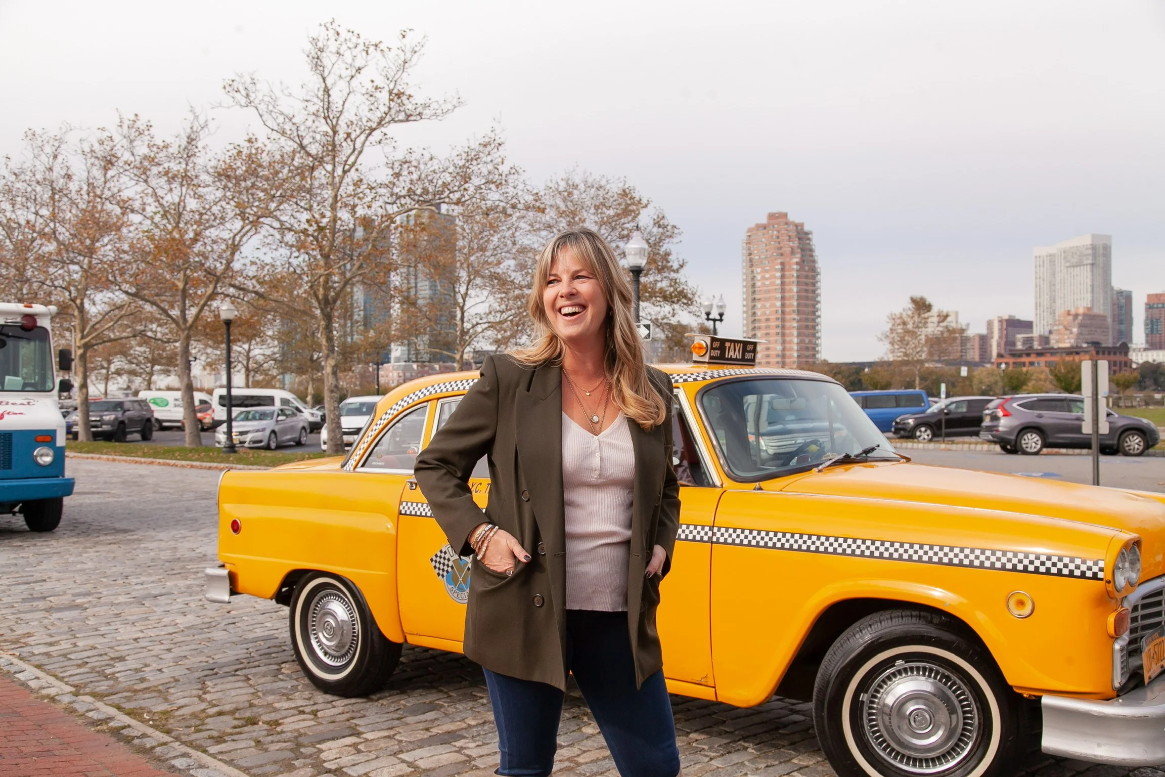 Nicole Darsney smiling wearing a green blazer and jeans standing next to a yellow taxi cab in Jersey Cityy with trees and tall buildings in the background.