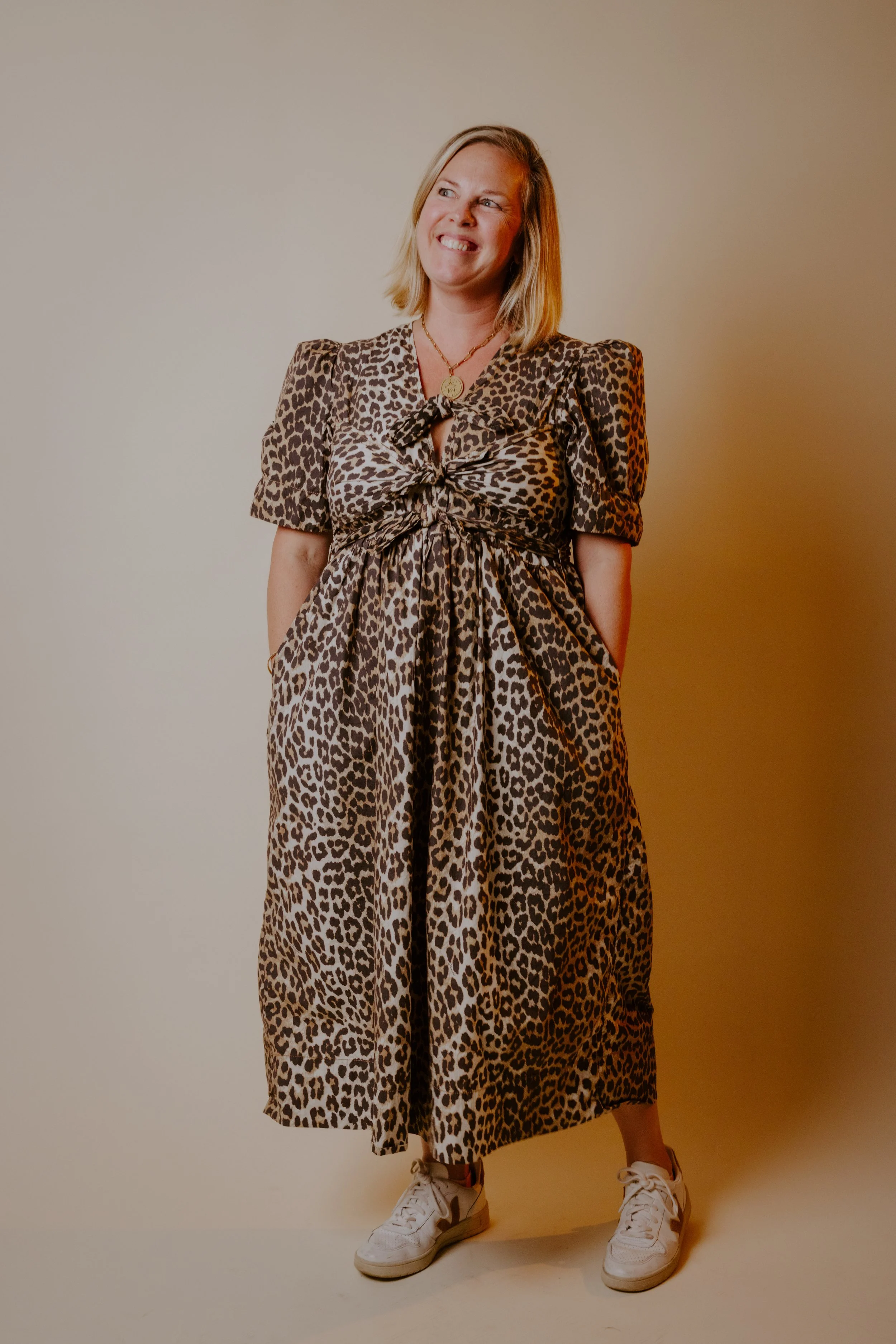 Nicole M. Darsney, a woman wearing a leopard print dress and white sneakers, standing against a plain background, smiling with her hands in her pockets.