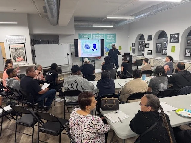 A classroom with a diverse group of adults attending a lecture. A speaker stands in front of a whiteboard and a large screen displaying a slide with charts. The room has framed photos on the walls, overhead ducts, and several tables and chairs.