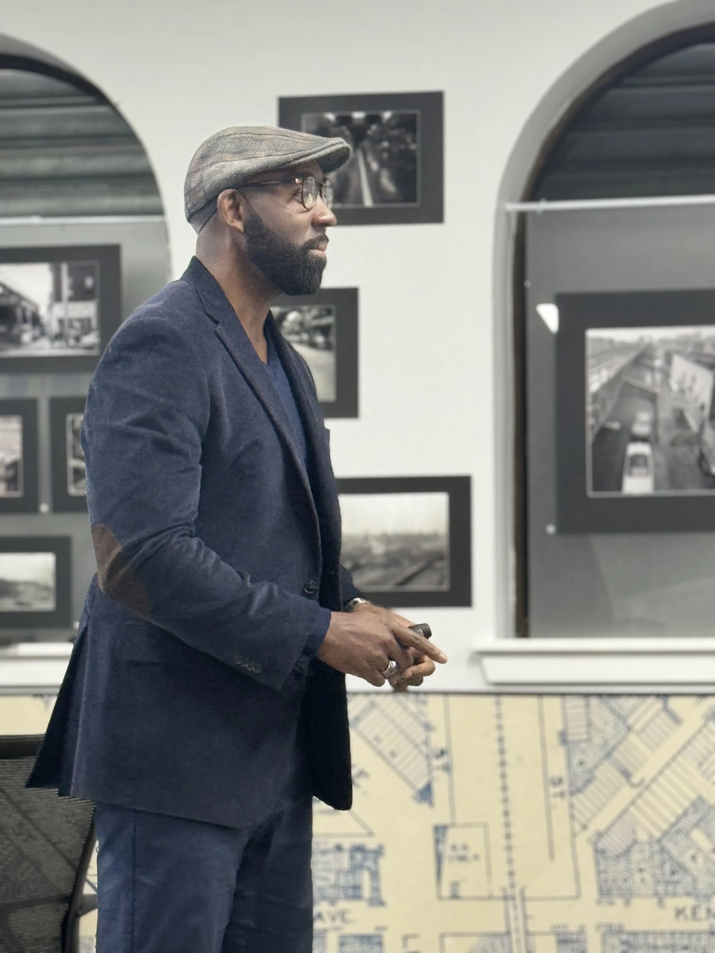 A man with a beard, glasses, and a flat cap, dressed in a navy blazer, standing with his hands clasped in front of him inside a room with black and white framed photographs on the wall and architectural plans on a table.