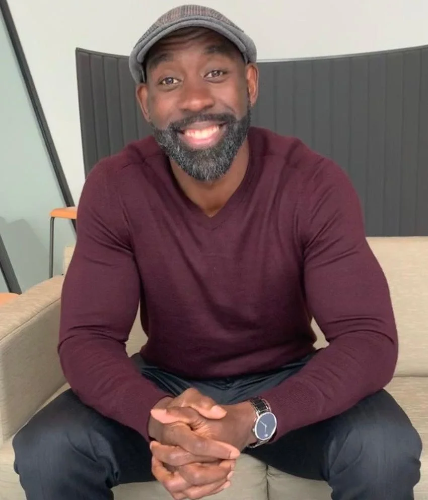 A smiling man with a beard, wearing a gray cap, maroon long-sleeve shirt, and watch, sitting on a beige couch.