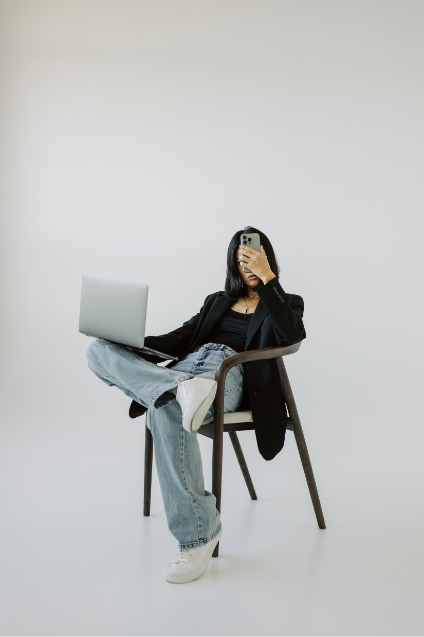 Young woman sitting on a chair with a laptop on her lap, holding a smartphone in front of her face, in a minimalist white space.