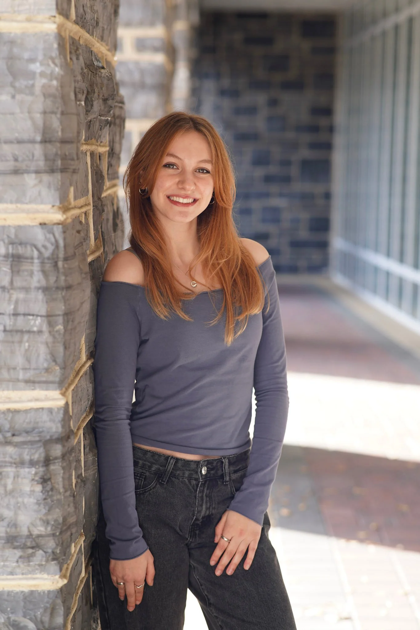 A young woman with red hair, wearing a gray off-the-shoulder top and black jeans, leaning against a stone wall on a bright day.