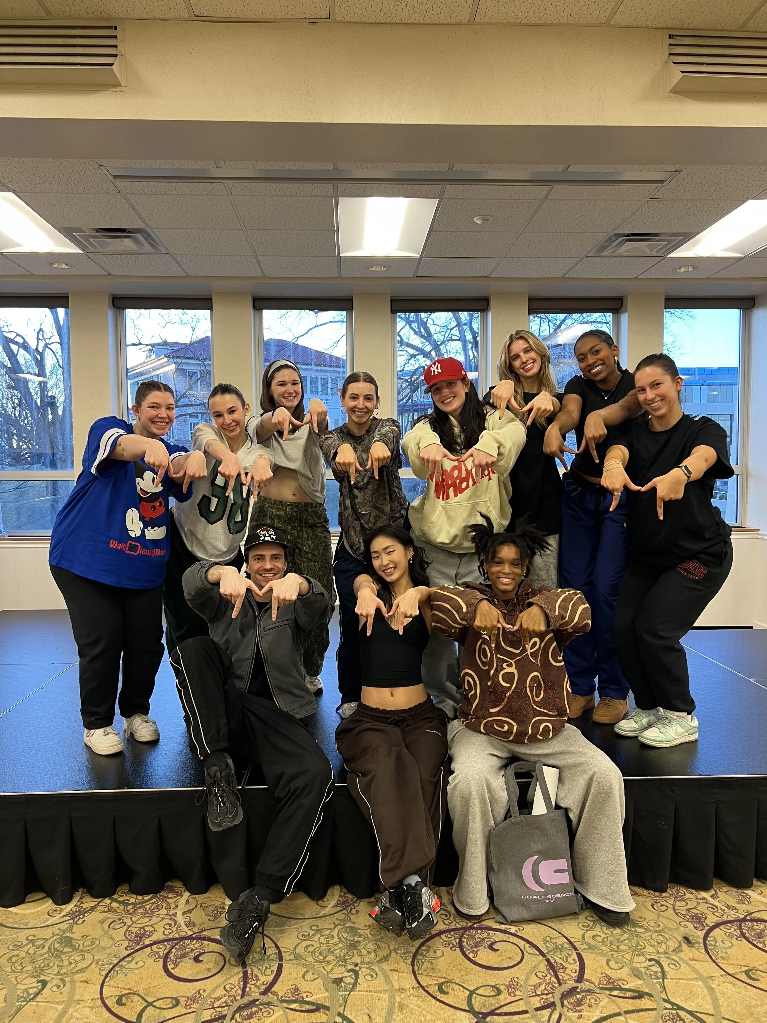 A group of twelve young dancers in casual and dancewear posing together in a rehearsal studio. They are pointing their fingers forward, smiling, with a large window behind them showing outdoor trees and buildings.