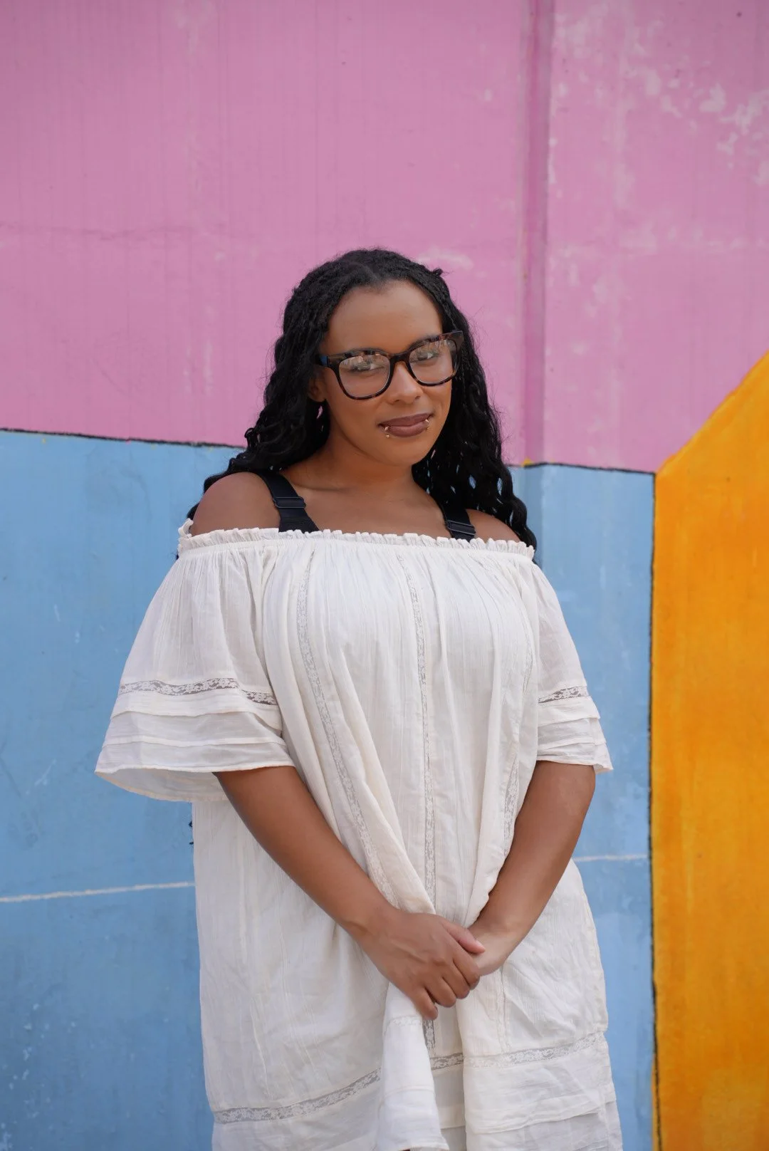 A young woman with curly hair smiling and standing against a stone wall in a casual outfit with a black top and beige denim skirt.