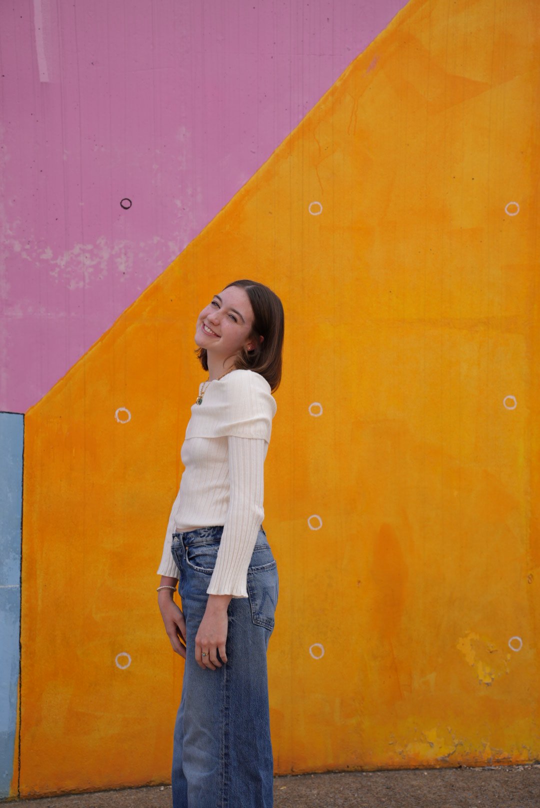 A young woman with long wavy hair and a bright smile standing outdoors, wearing a black long-sleeve top, a leopard print vest, and light gray jeans, with a modern building and glass windows in the background.