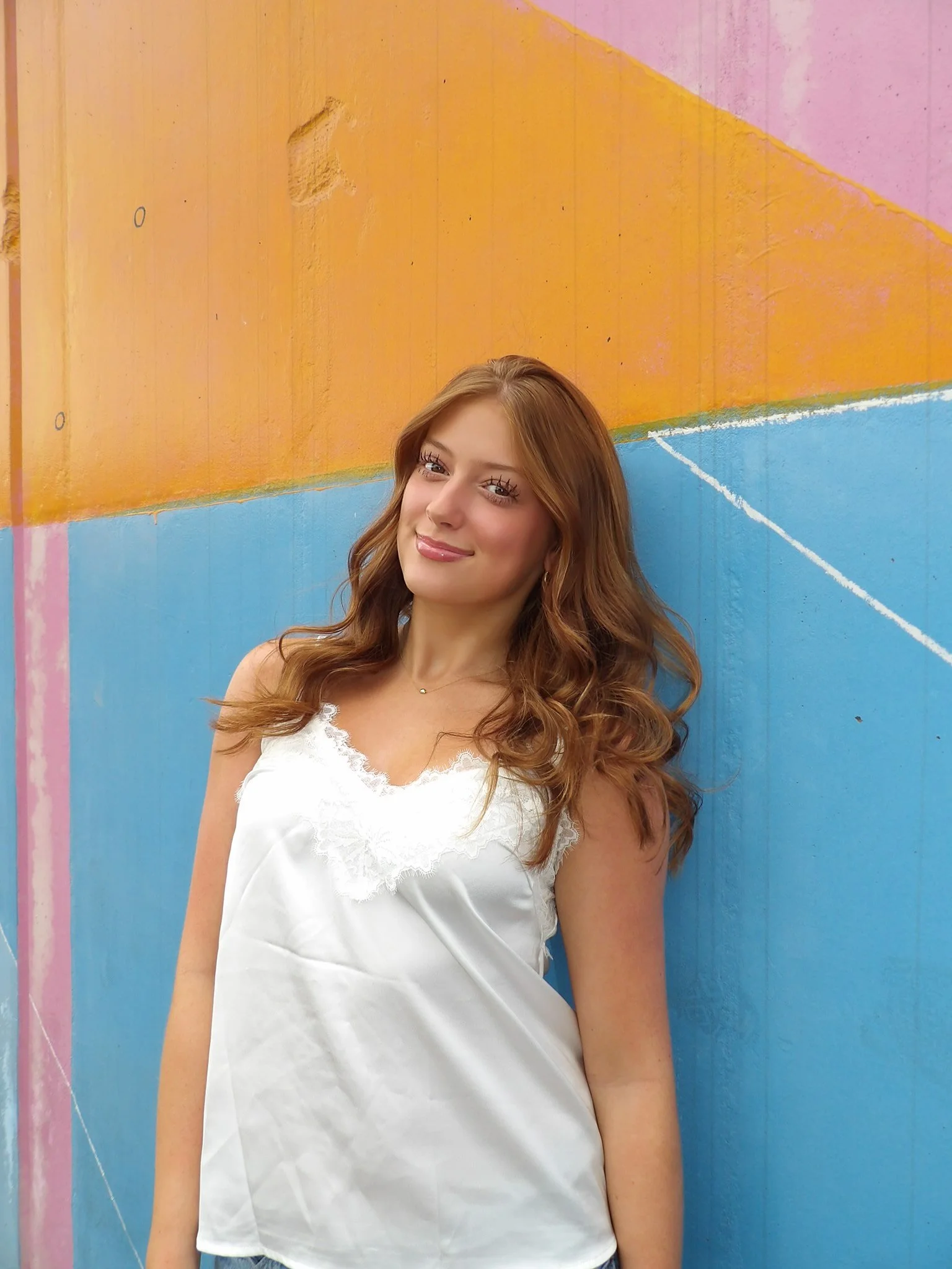 A young woman with shoulder-length brown hair, blue eyes, and light skin leaning against a stone wall, smiling at the camera. She is wearing a black off-the-shoulder top, a blue denim jacket, and matching jeans, with gold hoop earrings.