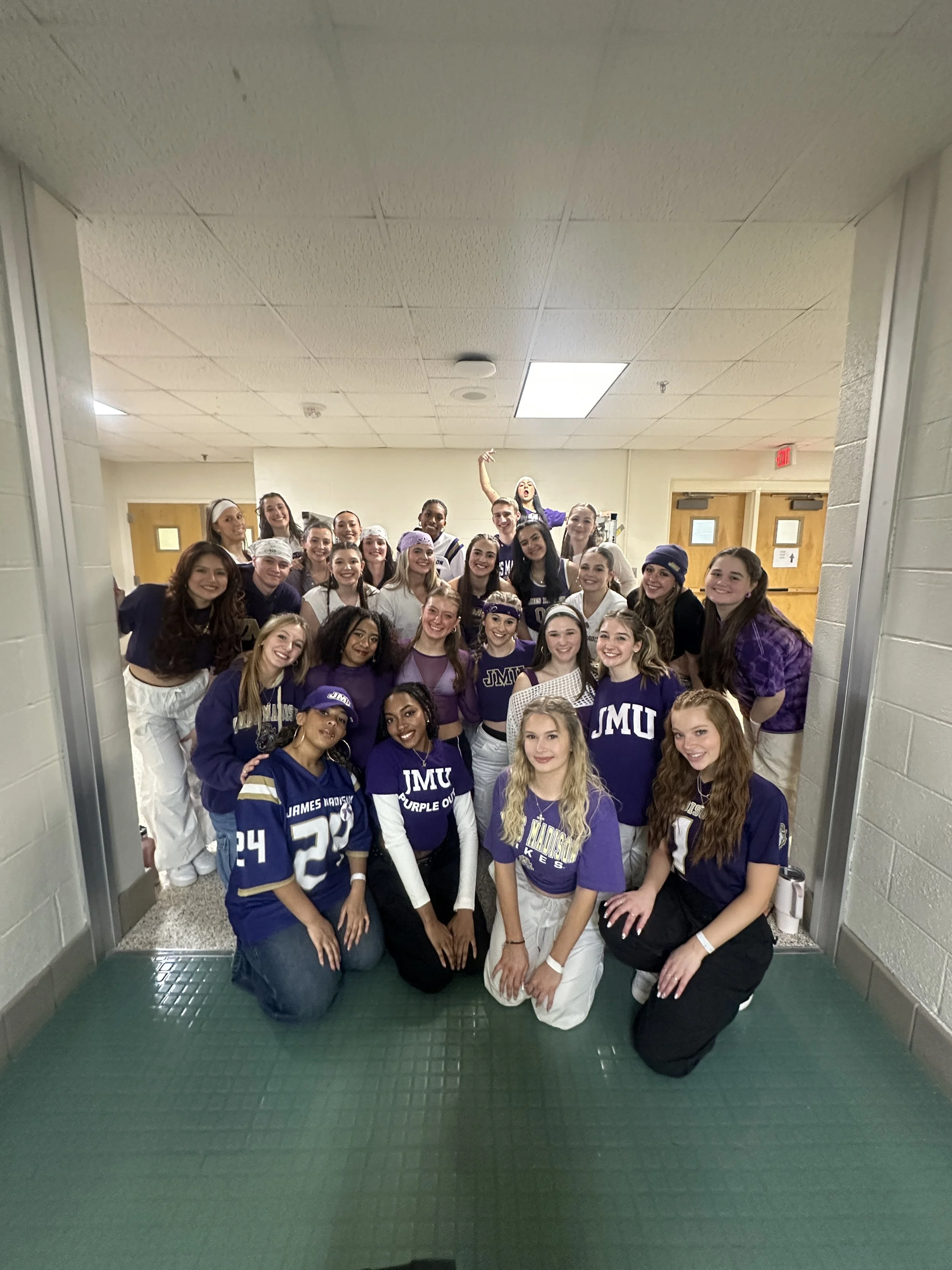 Group of women in purple and white sports uniforms and casual clothing posing together in a hallway, smiling and cheering.