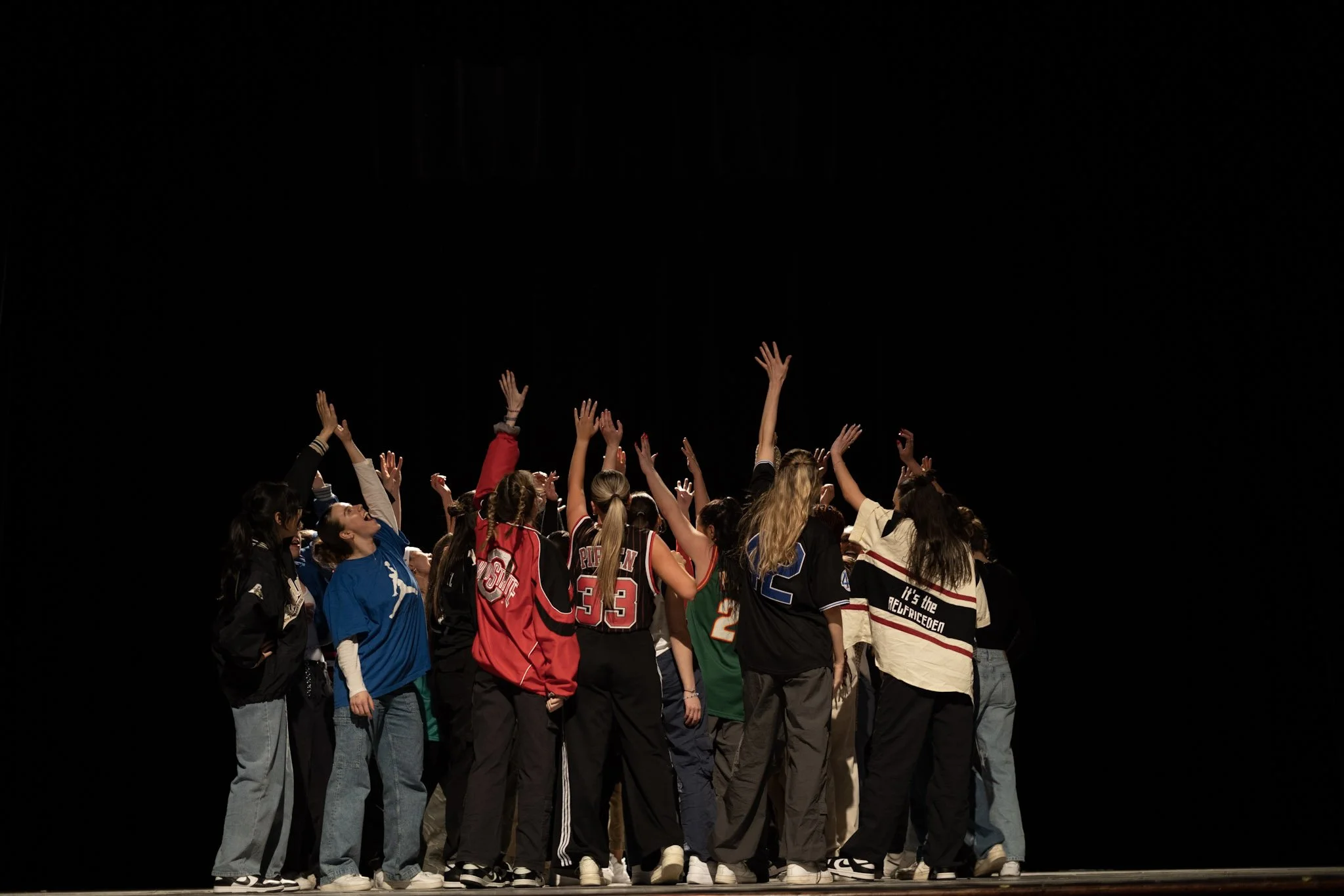 A group of young women and girls standing on a stage, raising their hands, facing upward, in a dark background.