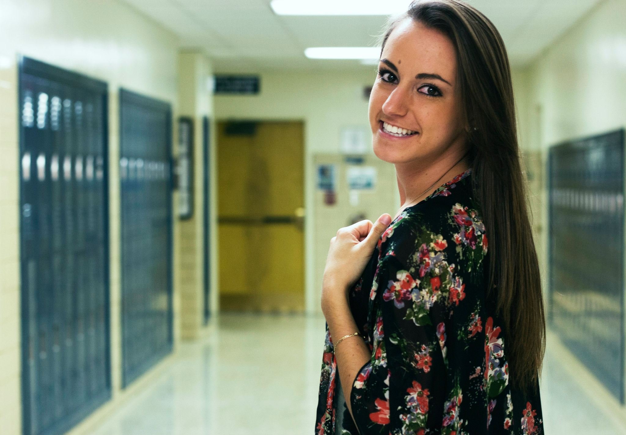 A smiling young woman with long brown hair standing in a school hallway next to lockers, wearing a floral-patterned jacket.