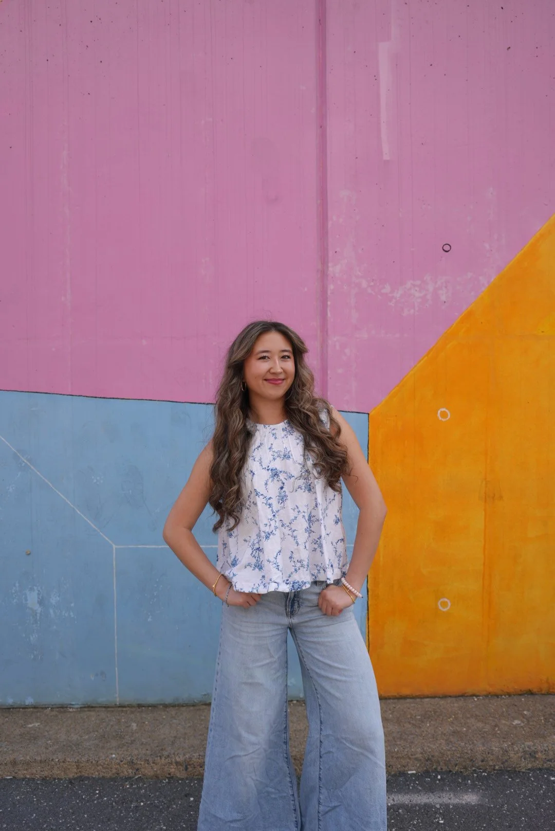 A young woman with red hair, wearing a gray off-the-shoulder top and black jeans, leaning against a stone wall on a bright day.