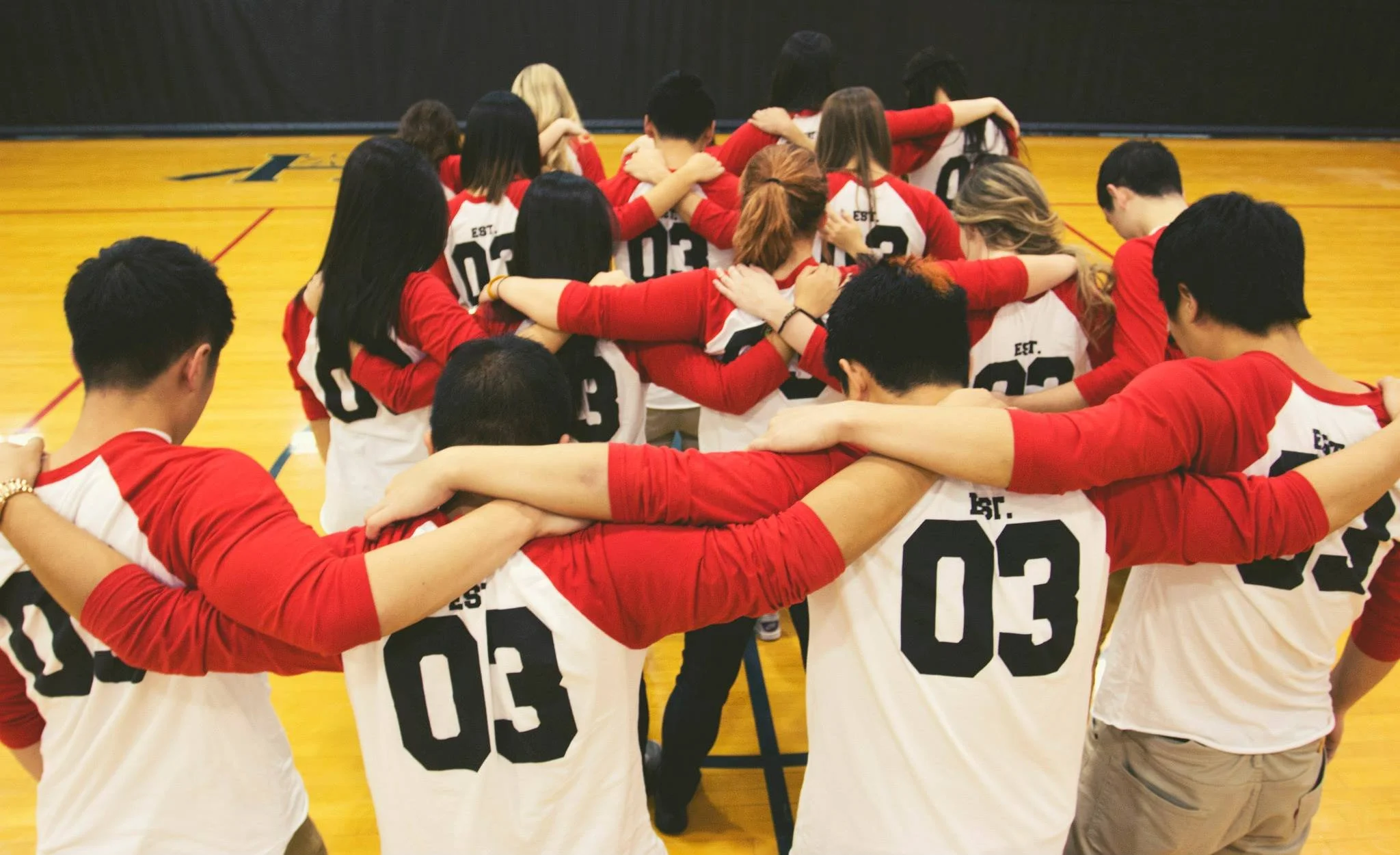 Group of people in red and white sports jerseys with their arms around each other's shoulders, standing in a circle on a basketball court.