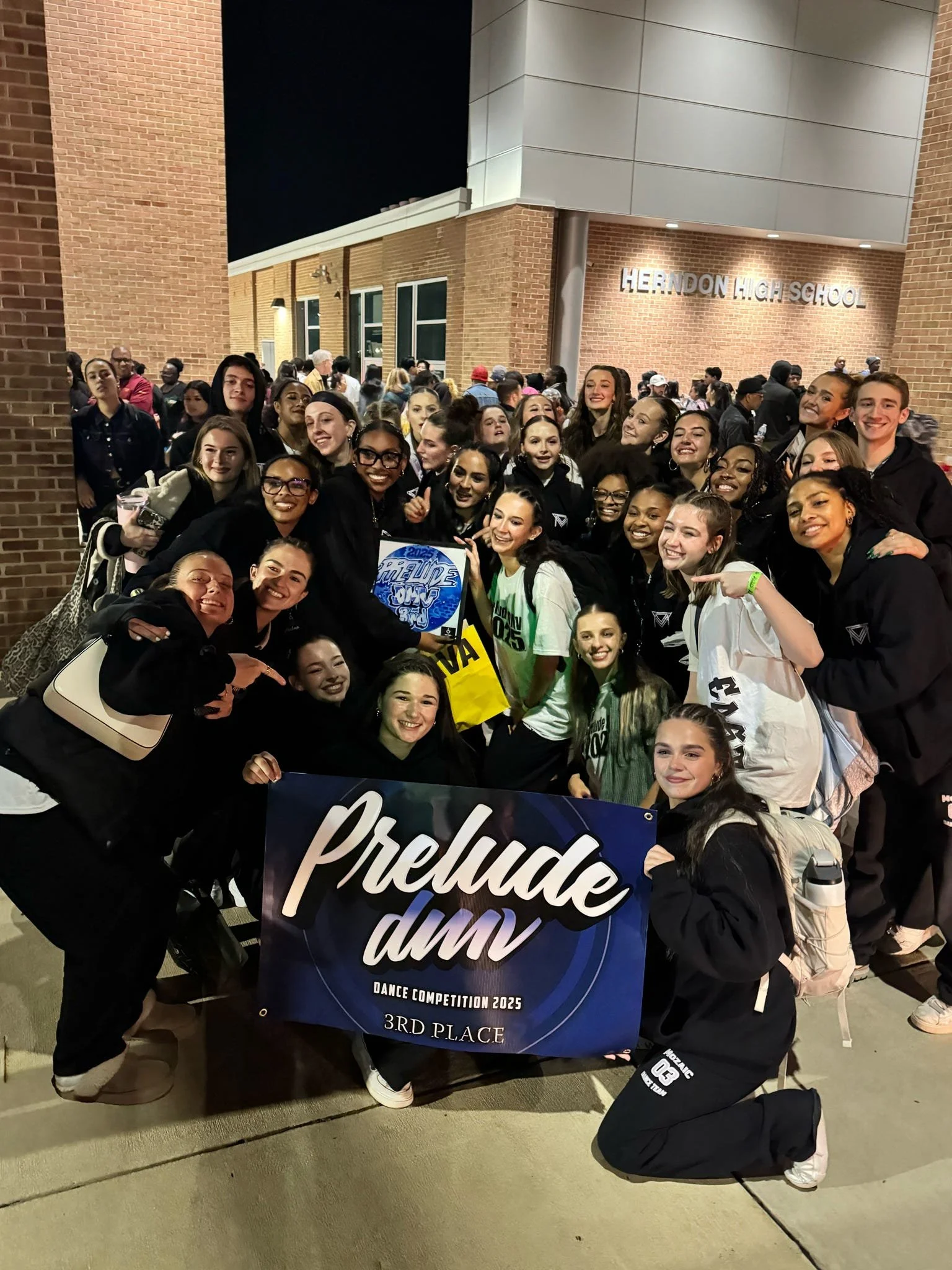 Group of cheerleaders at Herndon High School, holding a banner that reads 'Prelude dmv Dance Competition 2025 3rd Place,' posing outside with many spectators in the background.