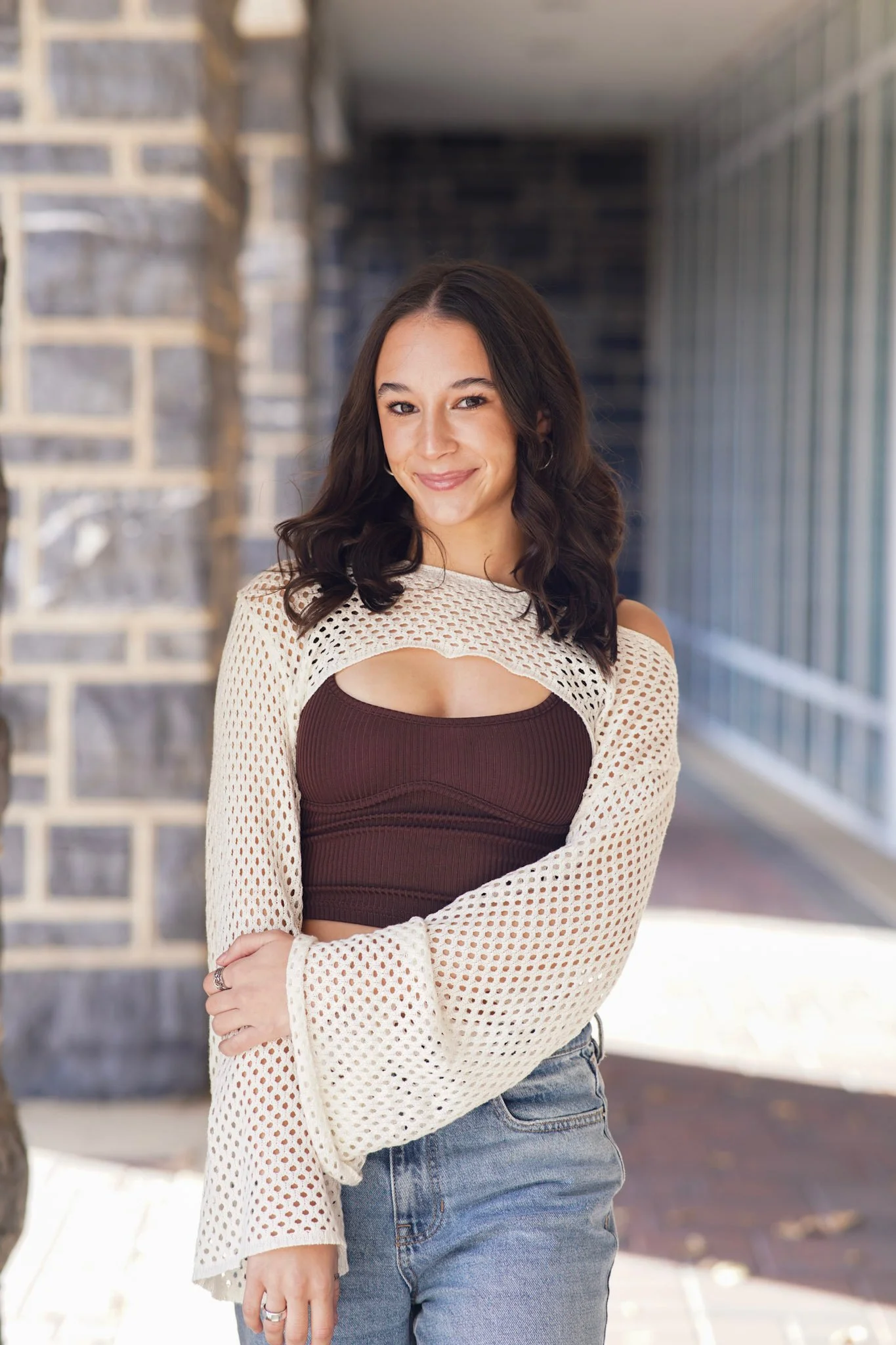 A young woman with long dark hair and hoop earrings standing outdoors in front of a stone wall and glass structure, smiling with her arms crossed, dressed in a brown top and a cream open-knit cardigan.