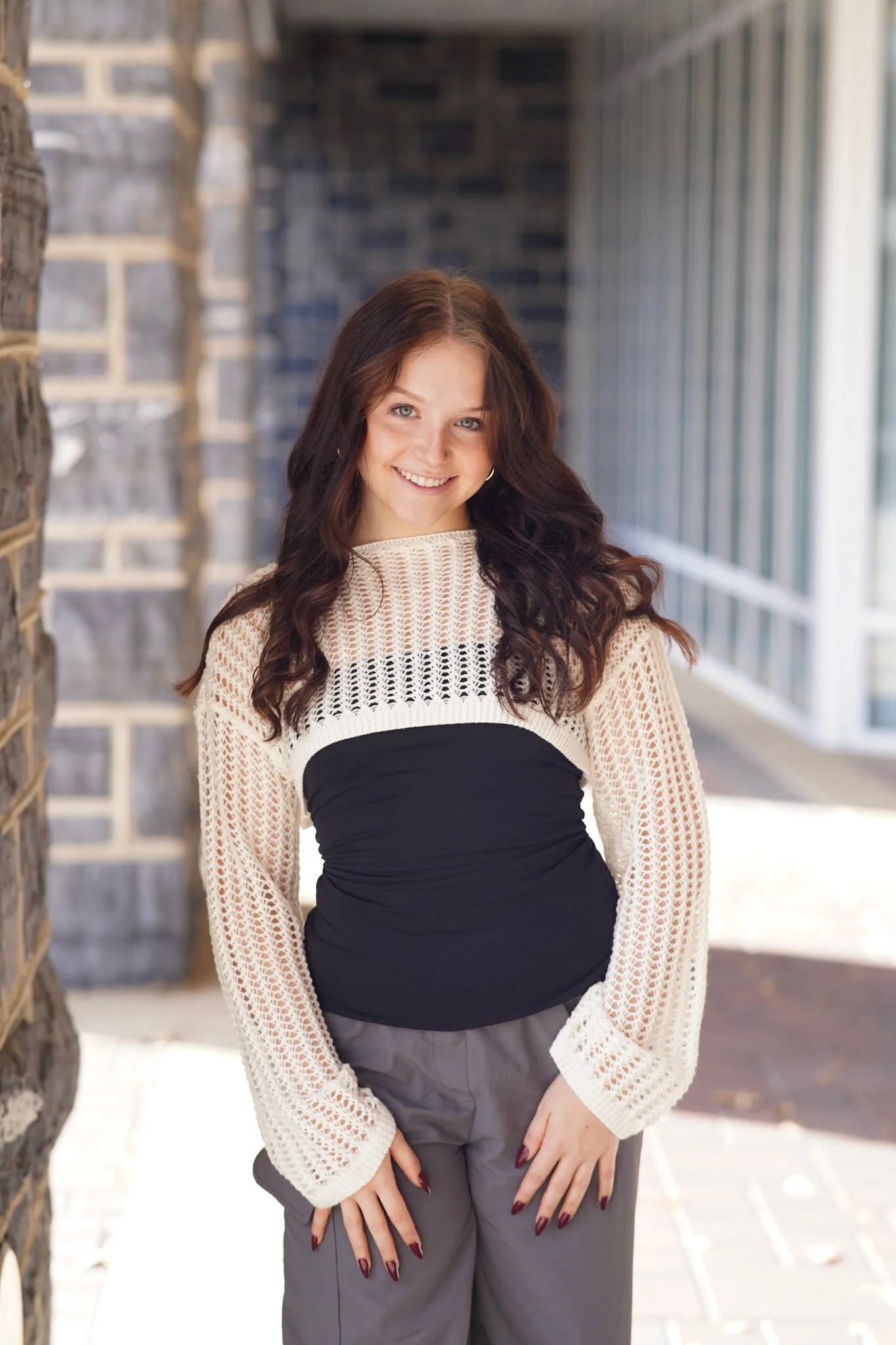 A young woman with dark brown, wavy hair, smiling, wearing a white crocheted top with long sleeves, a black bandeau, and gray pants, standing outdoors near a brick wall and glass windows.