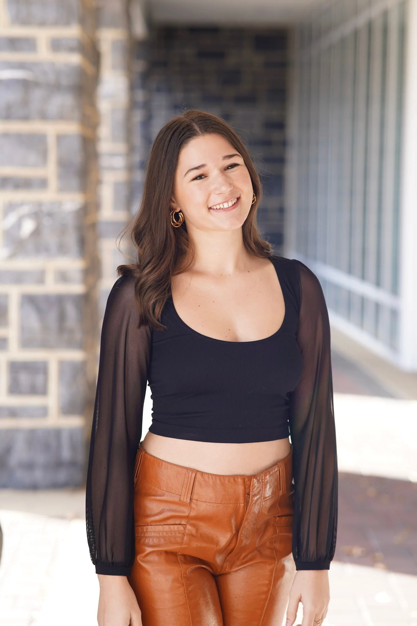 A young woman smiling outdoors, wearing a black crop top with sheer long sleeves and brown leather pants, standing in front of a brick wall and large window.