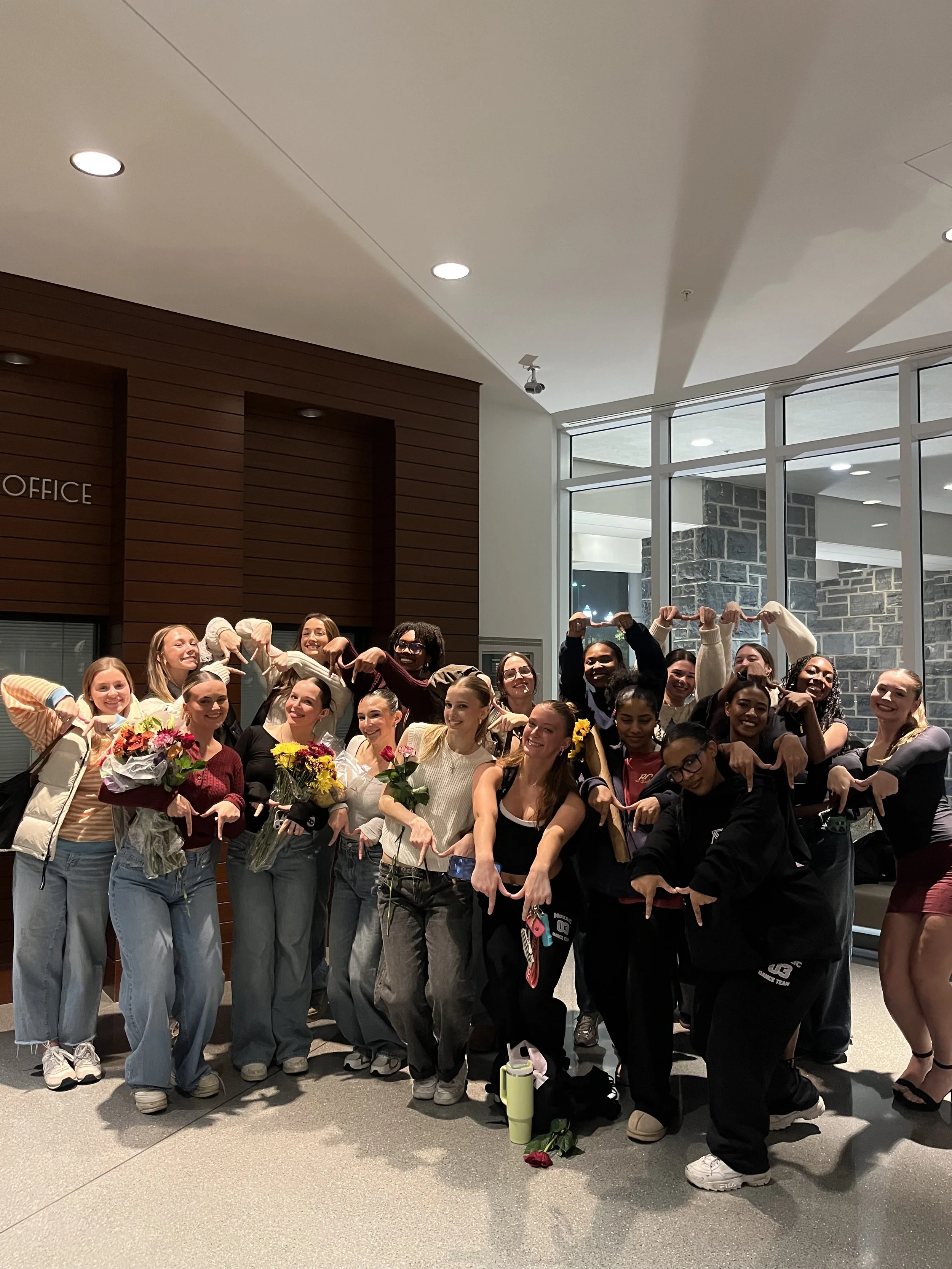 Group of women posing together indoors, some holding flowers, others making playful hand gestures, with large windows and a wooden wall in the background.
