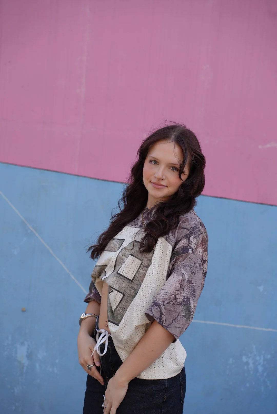 A young woman with long dark hair and hoop earrings standing outdoors in front of a stone wall and glass structure, smiling with her arms crossed, dressed in a brown top and a cream open-knit cardigan.