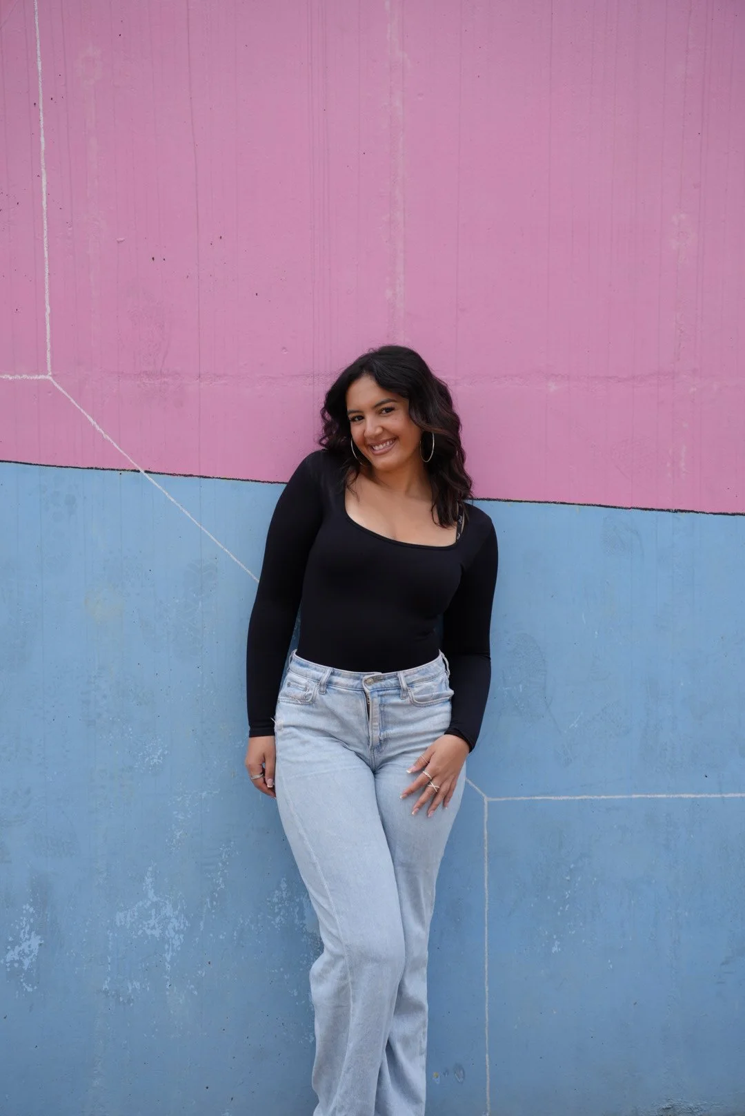 A young woman smiling outdoors, wearing a black crop top with sheer long sleeves and brown leather pants, standing in front of a brick wall and large window.