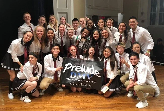 Group of young dancers in white shirts and dark skirts or pants, holding a banner that says 'Prelude DMV Urban Dance Competition 2018 2nd Place,' celebrating their win at the dance competition.
