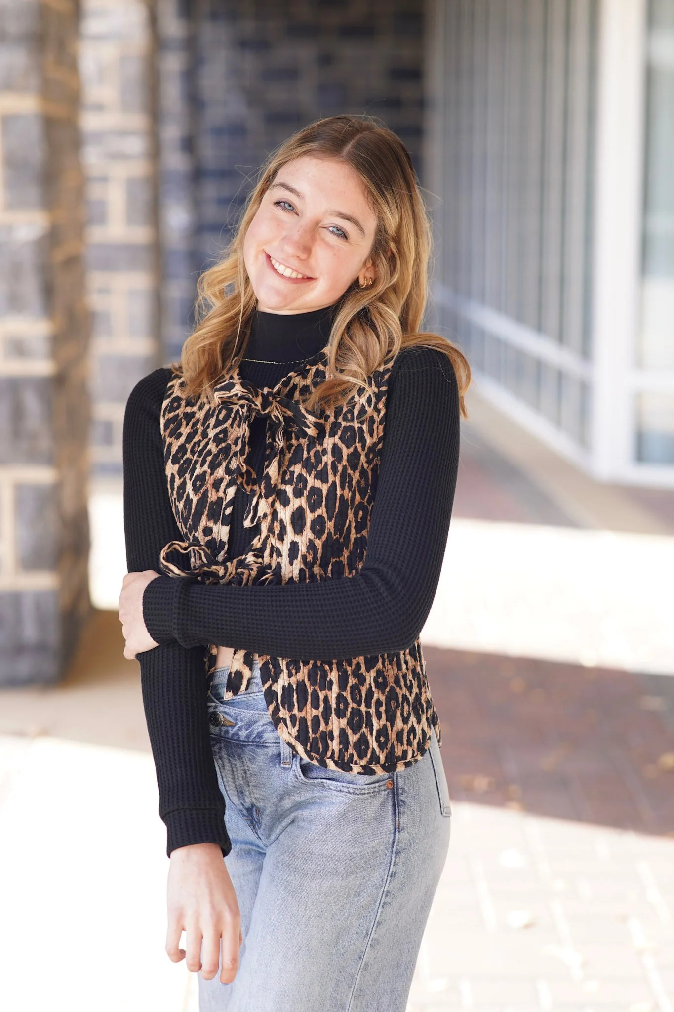 A young woman with long wavy hair and a bright smile standing outdoors, wearing a black long-sleeve top, a leopard print vest, and light gray jeans, with a modern building and glass windows in the background.
