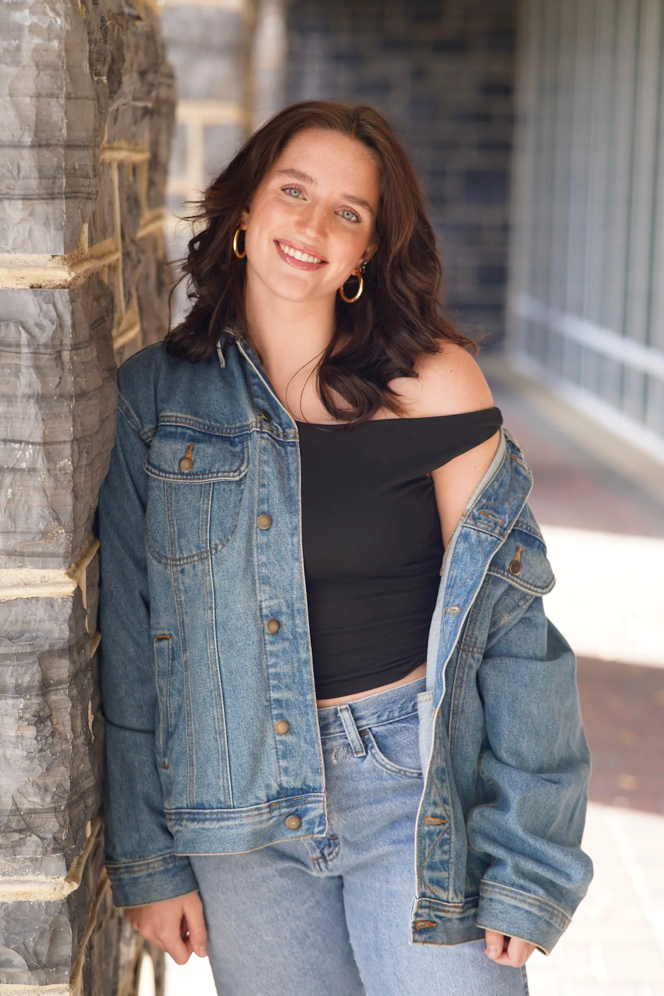A young woman with shoulder-length brown hair, blue eyes, and light skin leaning against a stone wall, smiling at the camera. She is wearing a black off-the-shoulder top, a blue denim jacket, and matching jeans, with gold hoop earrings.