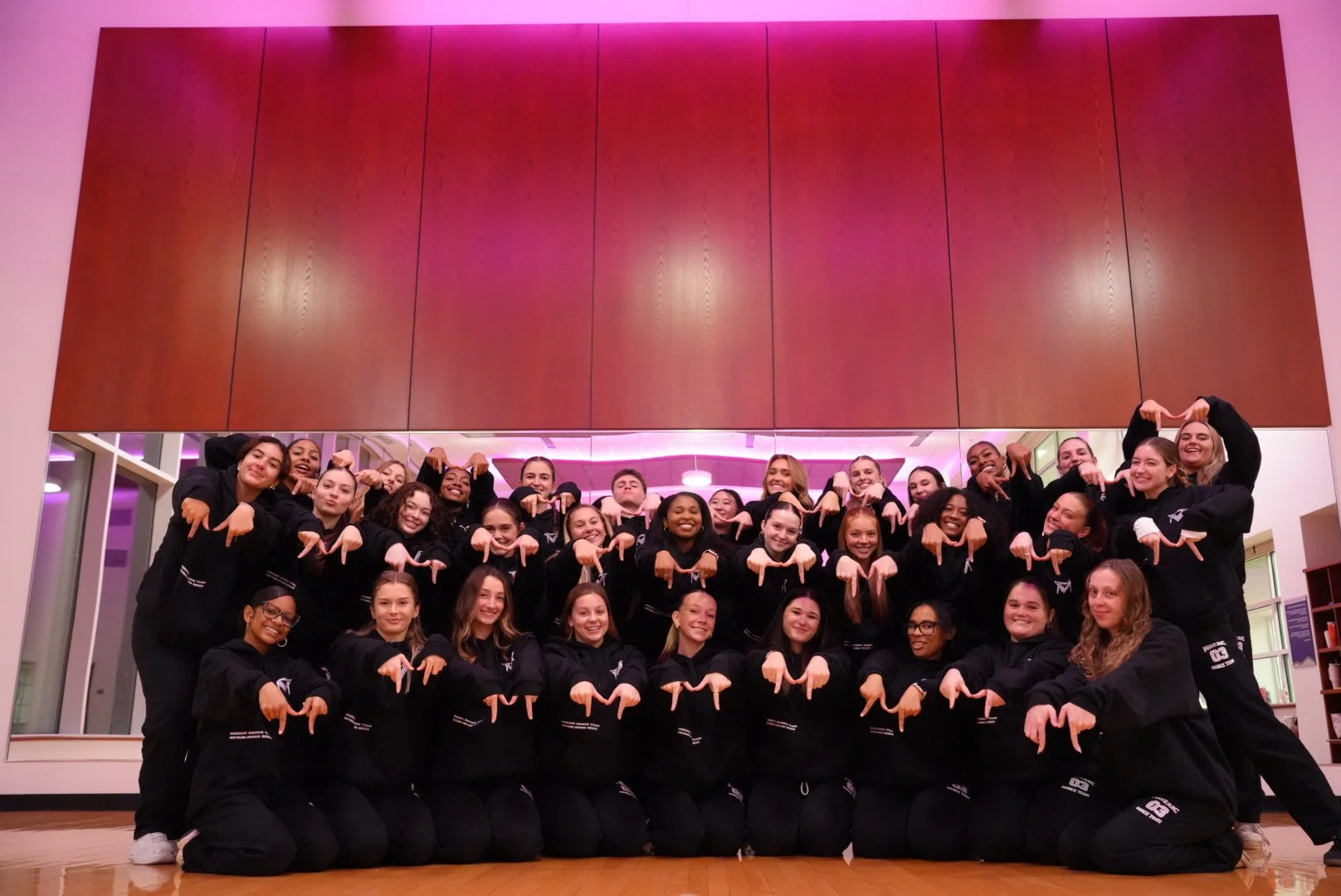 Group of young women dressed in matching black sweatshirts and pants, posing indoors with smiles and pointing downward with their index fingers.