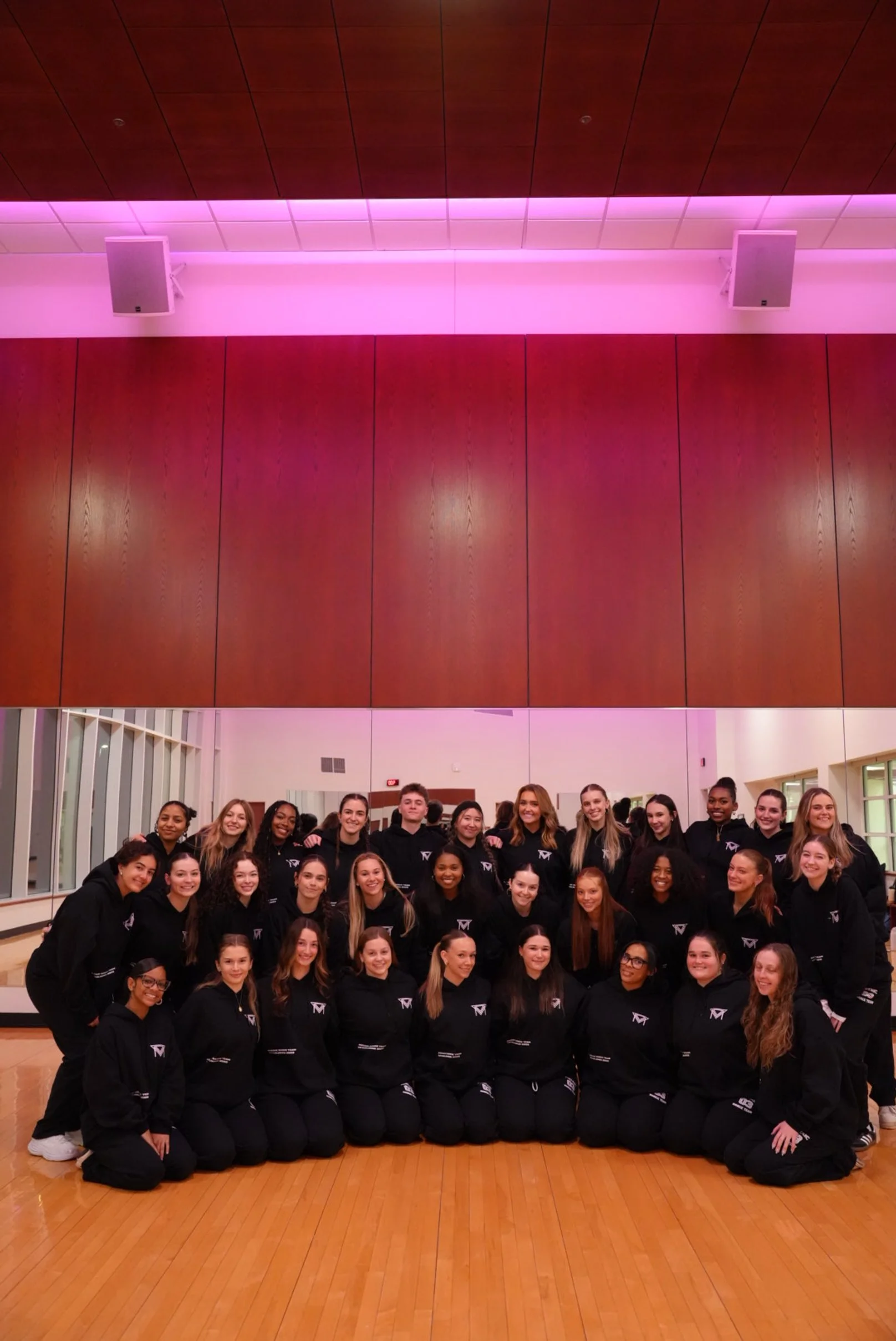 Group of young women in black hoodies posing for a photo in a room with wooden floors and a mirrored wall.