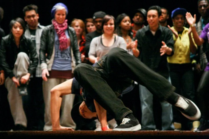 A breakdancer performing a headspin move on stage, surrounded by a diverse crowd of onlookers watching and clapping.