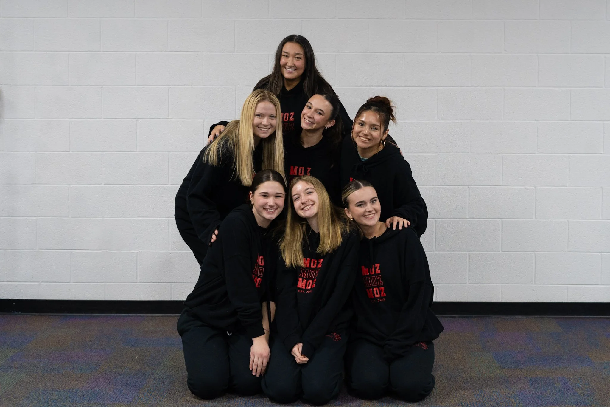Group of eight young women wearing matching black hoodies with red text, smiling and posing together against a white wall.
