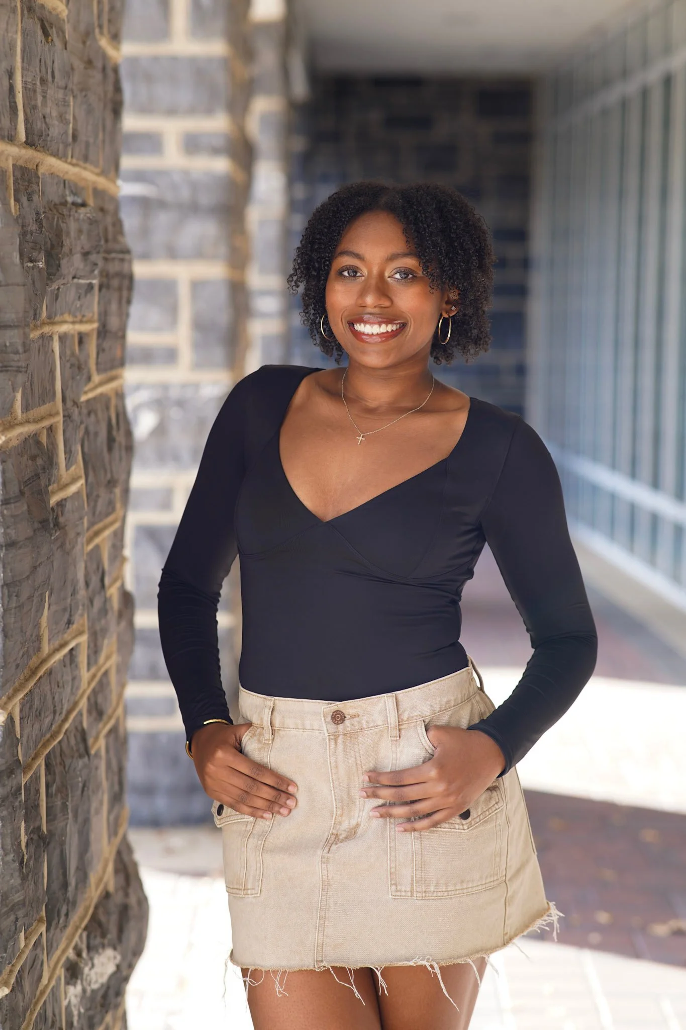A young woman with curly hair smiling and standing against a stone wall in a casual outfit with a black top and beige denim skirt.