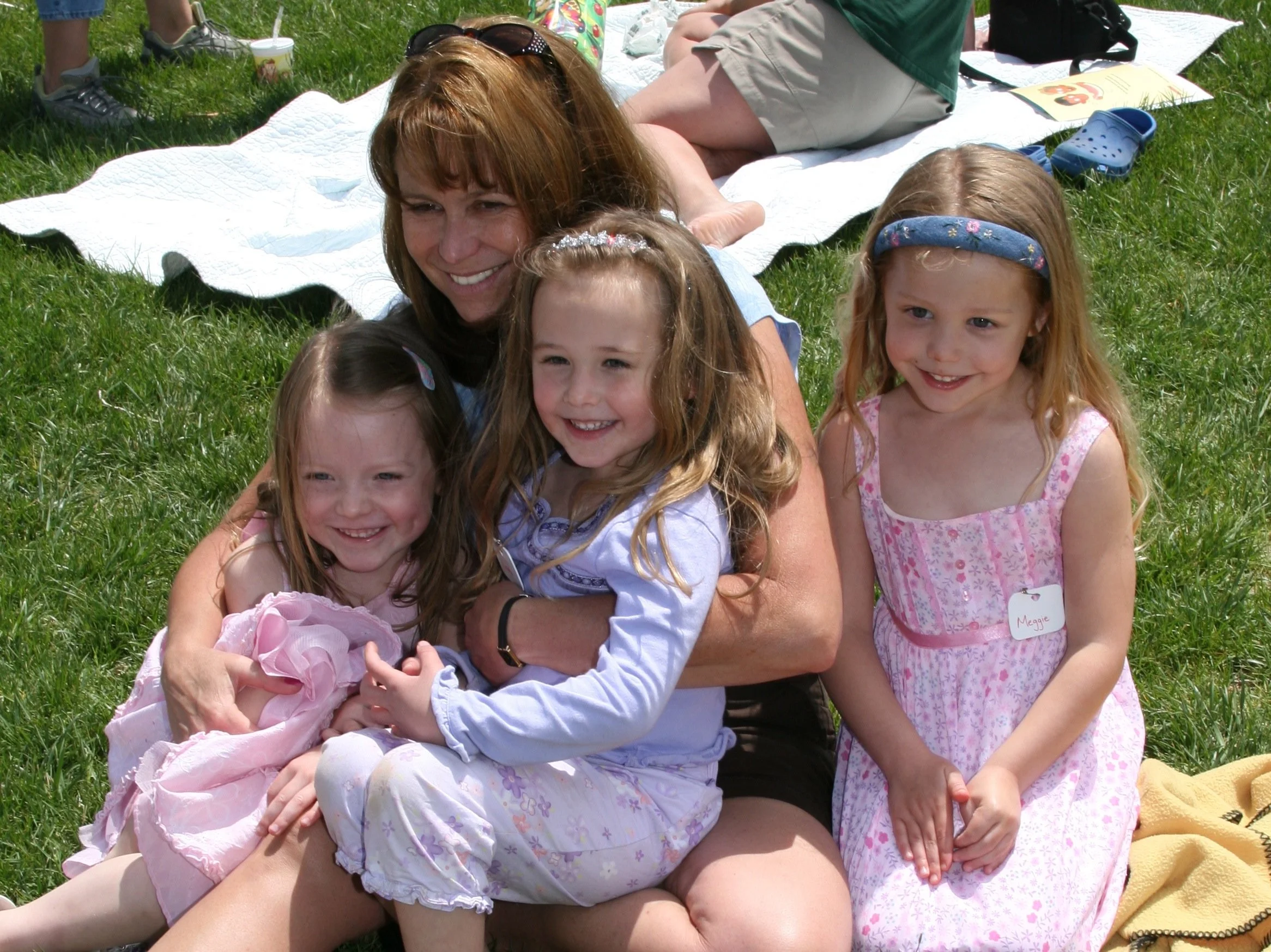 A woman surrounded by four young girls sitting on grass, smiling, with blankets and picnic items in the background on a sunny day.