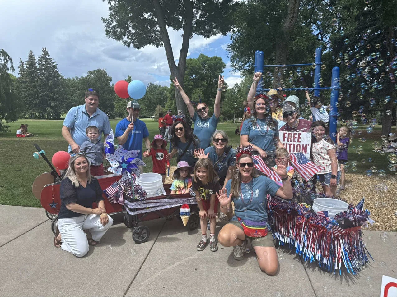 Group of people celebrating outdoors, holding American flags, balloons, and patriotic decorations, with a float decorated in red, white, and blue, featuring a sign that says "Free Education" and surrounded by bubbles.