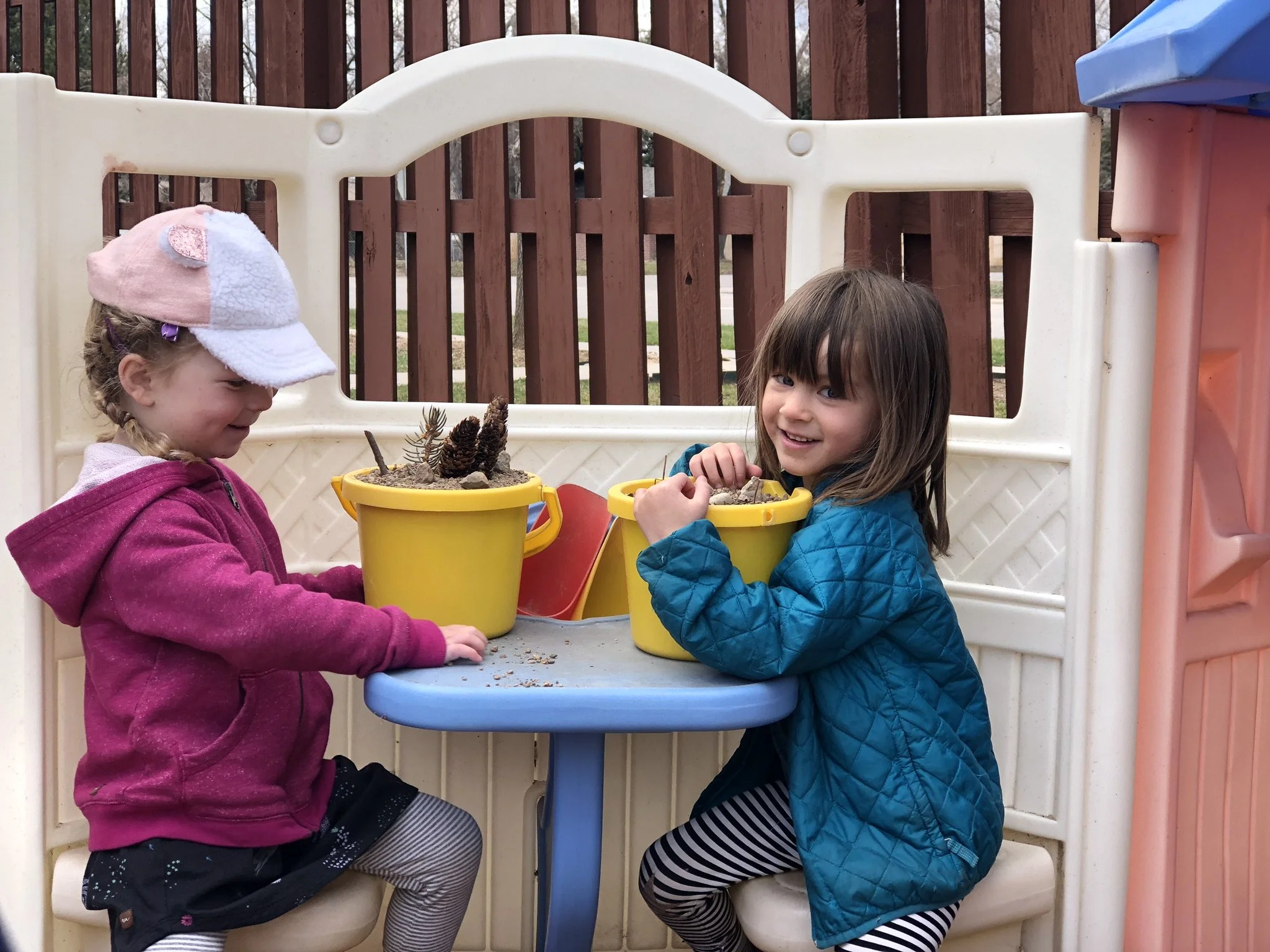Two young girls are sitting at a small blue table in a play area, smiling and playing with yellow buckets filled with dirt and pinecones.