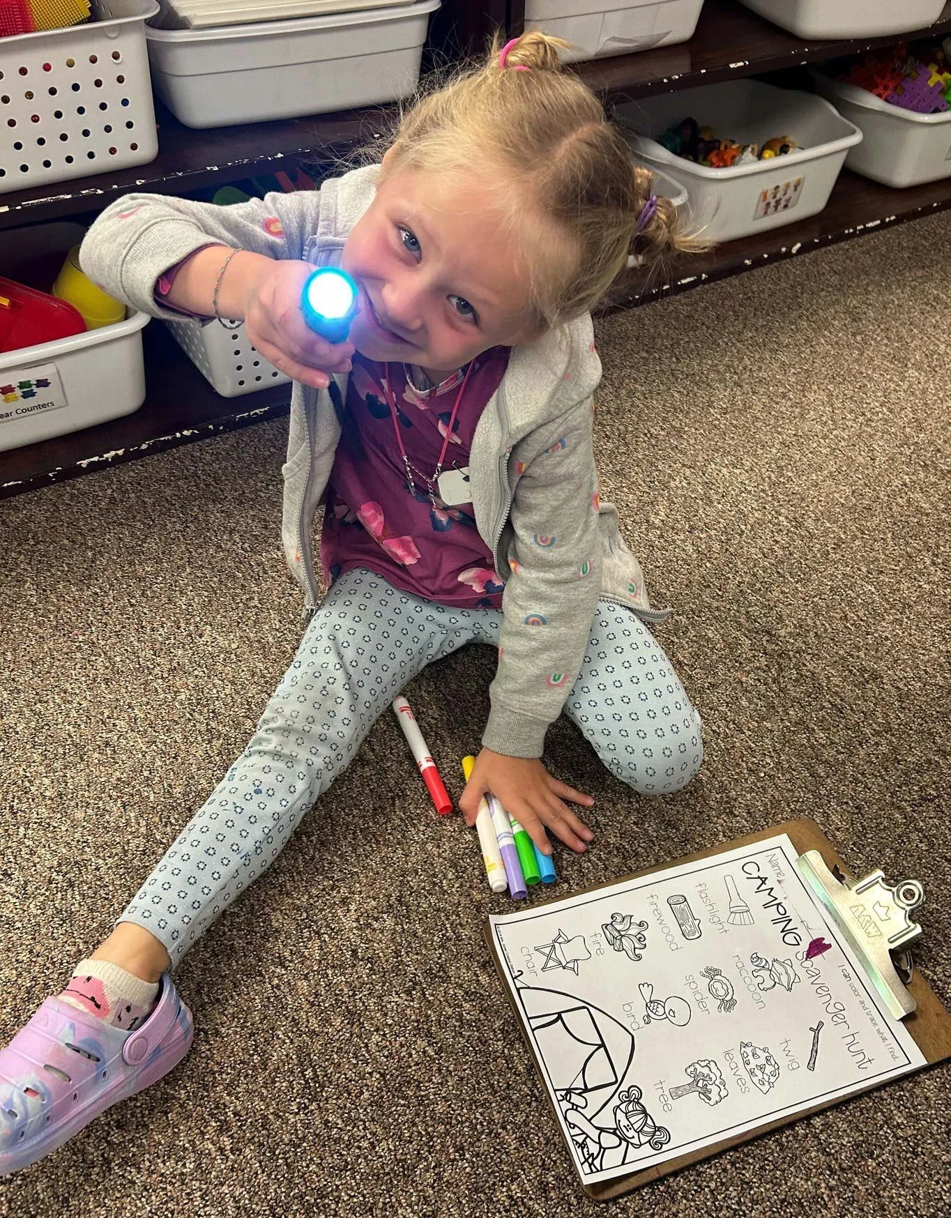 A young girl sitting on the carpeted floor, holding a flashlight pointed towards the camera. She is smiling and has colorful markers and a coloring sheet on a clipboard in front of her. The coloring sheet has a camping theme with illustrations of a tent, campfire, and outdoor activities.