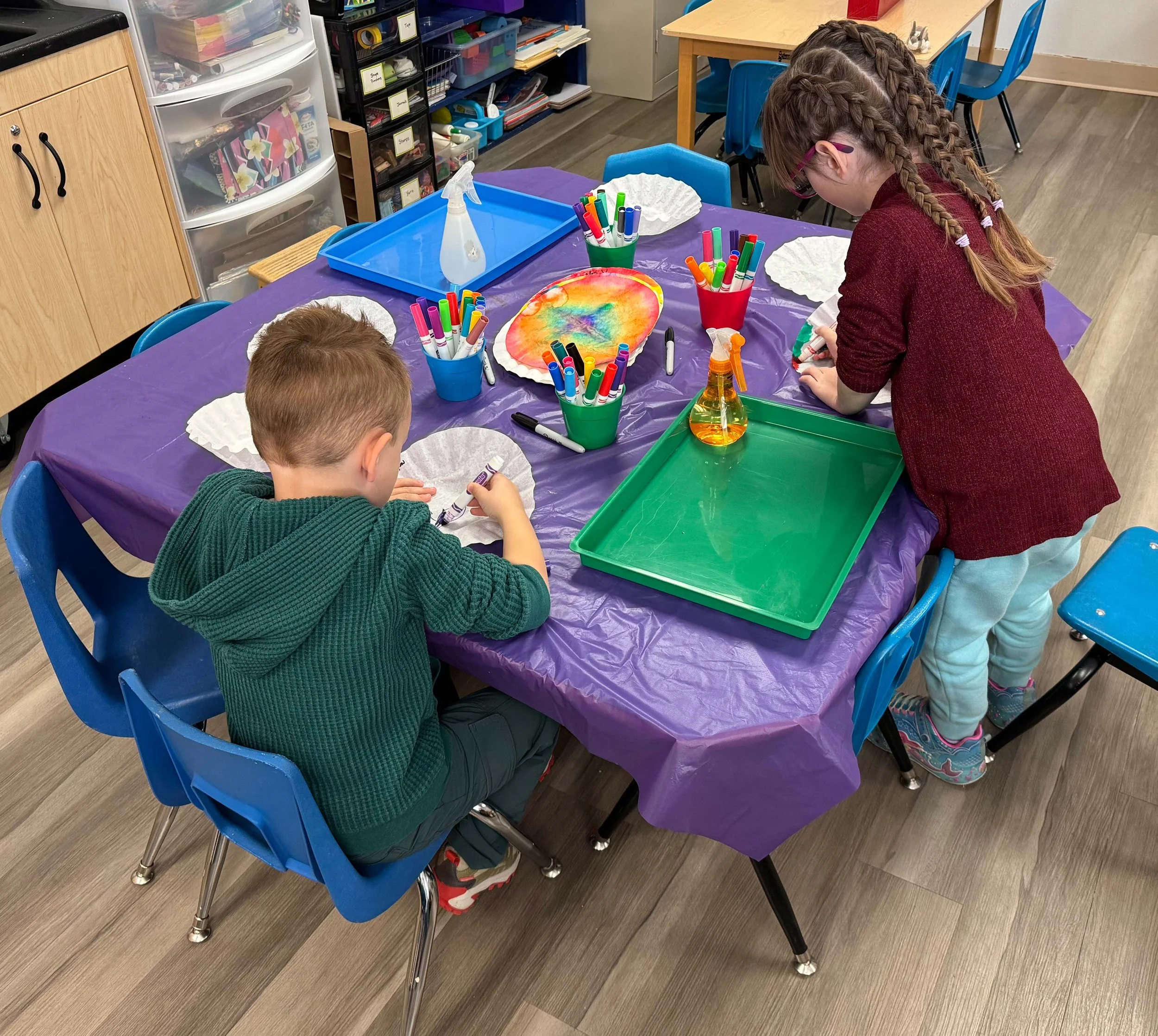 Two children creating tie-dye designs on white paper plates at a table in a classroom.