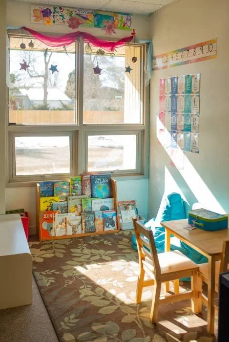 A brightly lit classroom corner with a window, book display, and educational posters on the wall.