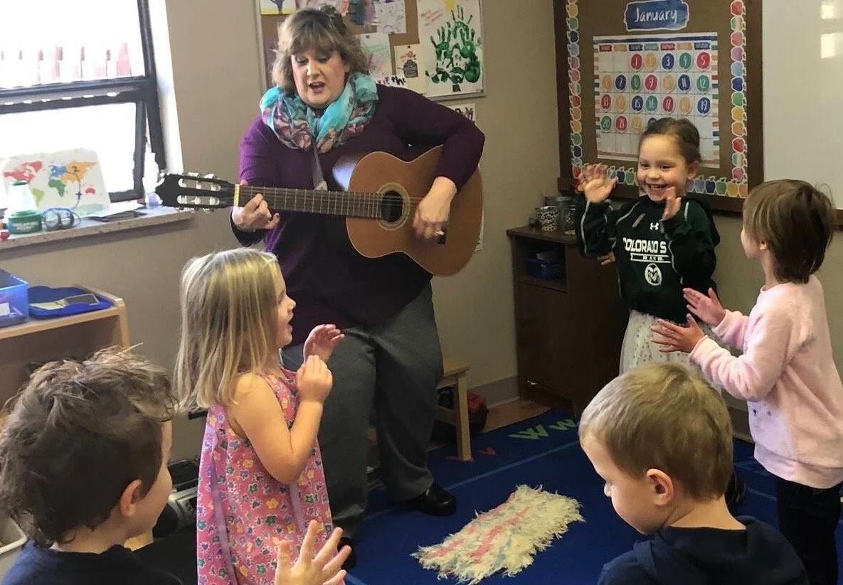 A woman is playing guitar and singing to young children in a classroom. The children are clapping and singing along, with some smiling and others engaging attentively.