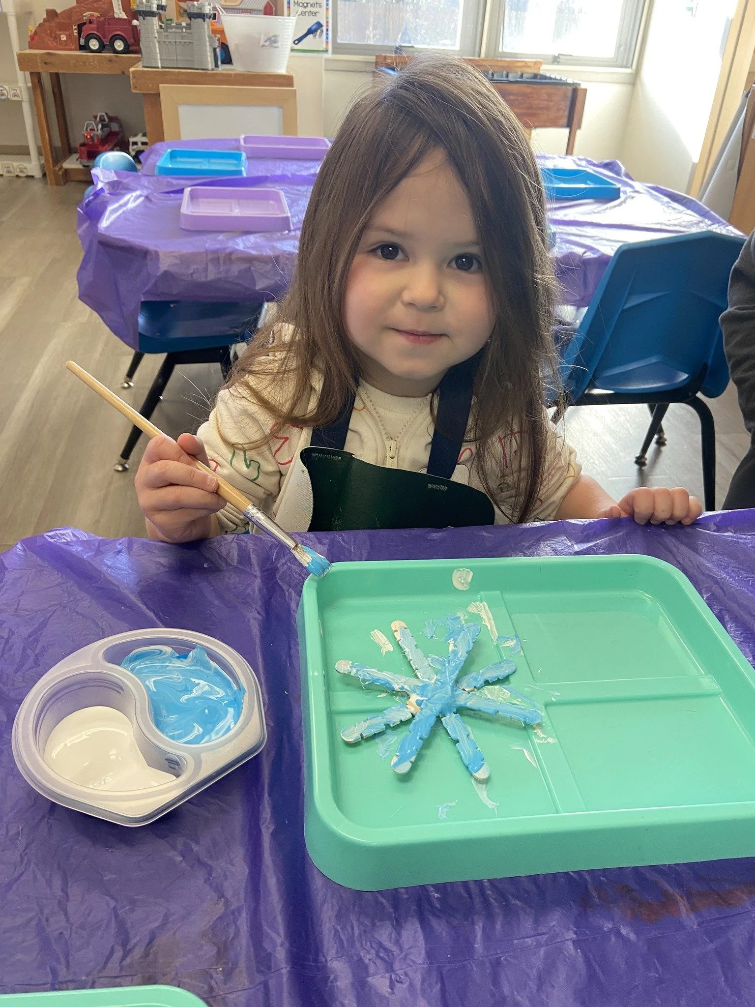 Young girl with brown hair holding a paintbrush at a craft table, creating a snowflake shape with blue paint on a green tray in a classroom setting.