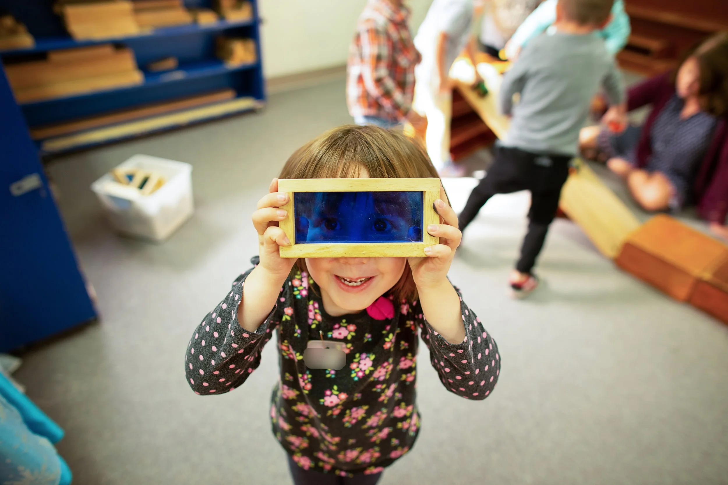 A young girl smiling and holding a small rectangular mirror with a wooden frame in front of her face, reflecting her eyes.