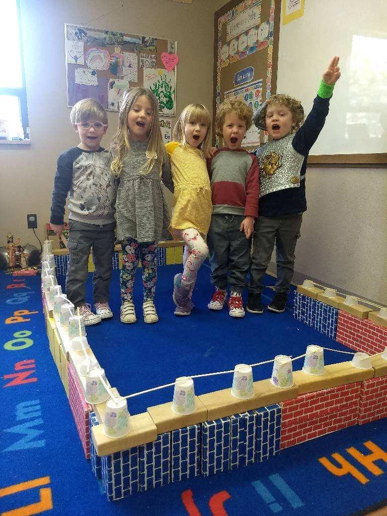 Five children standing on a small indoor play structure decorated with cups and paper cups, singing or talking.