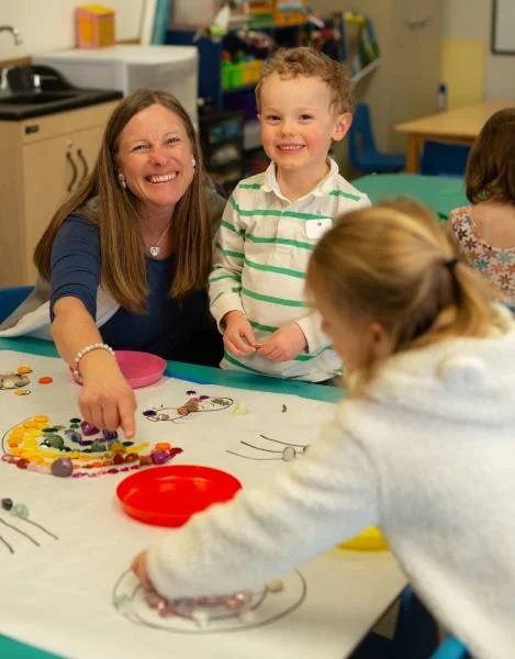 A woman and a young boy smiling at a classroom table with art supplies, while a girl with a ponytail is drawing on paper.