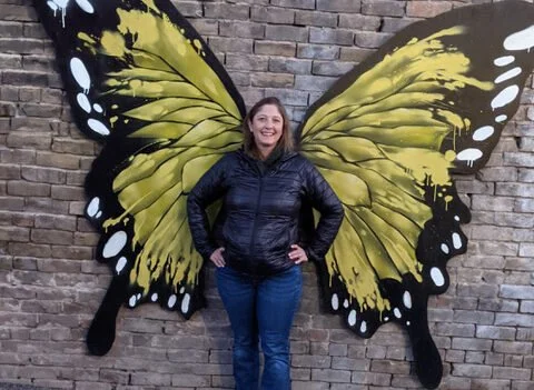 Woman standing in front of a mural painted on a brick wall, resembling butterfly wings with yellow and black patterns.