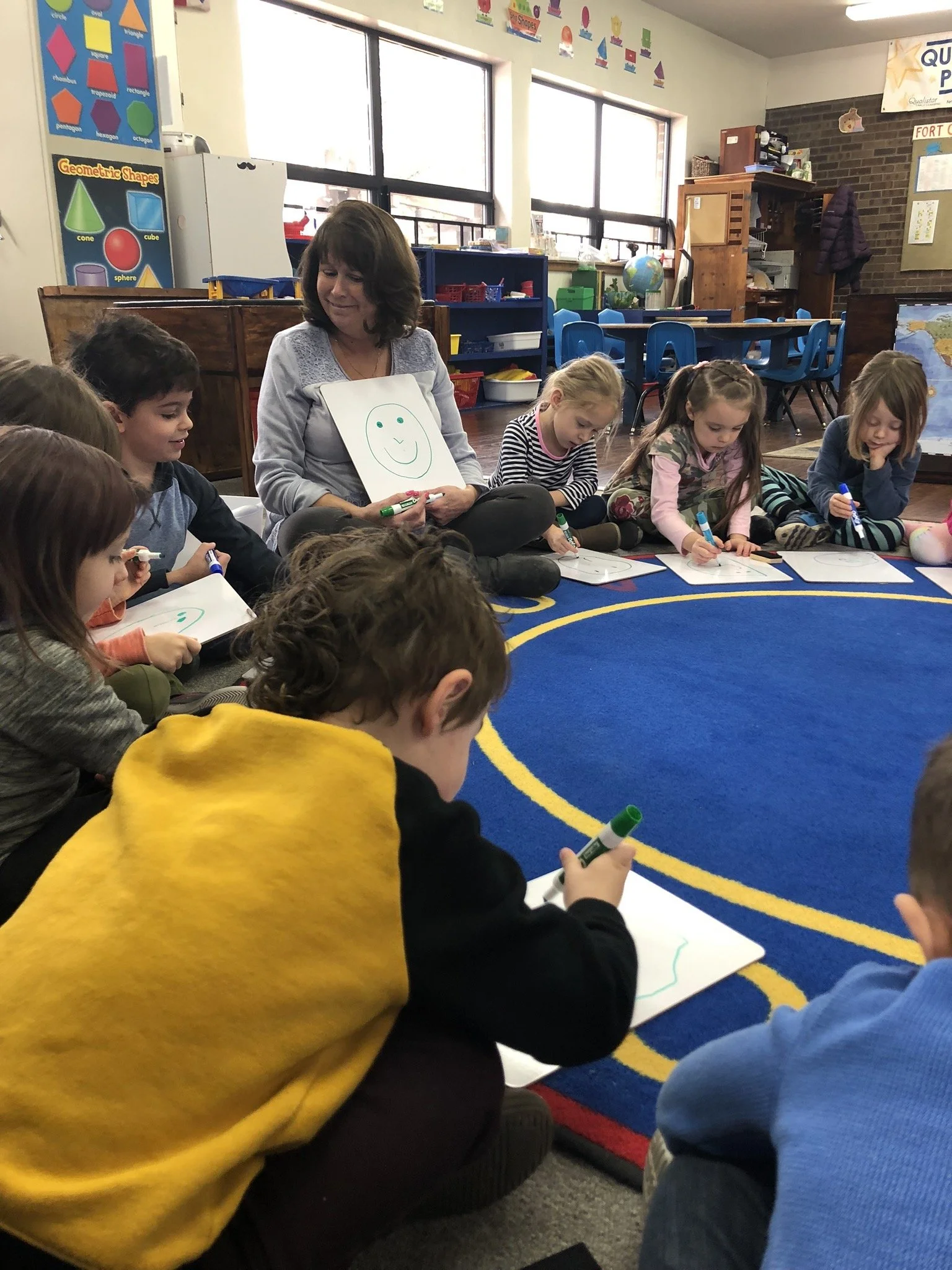 Kindergarten classroom with children sitting on a blue rug, drawing on whiteboards, and a teacher holding a whiteboard with a smiley face.