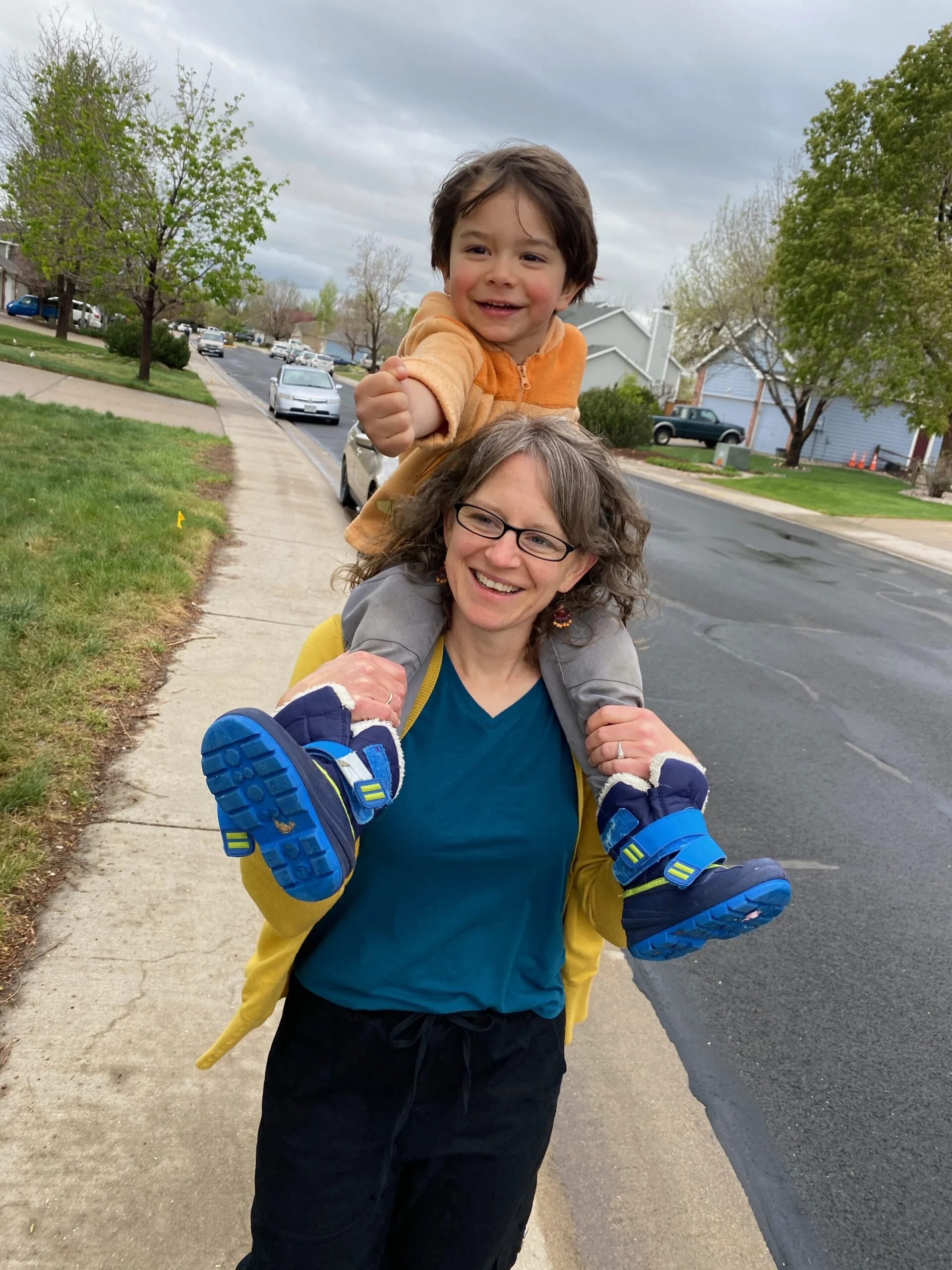 A woman carrying a young boy on her shoulders walking on a sidewalk in a neighborhood, with trees and houses in the background.