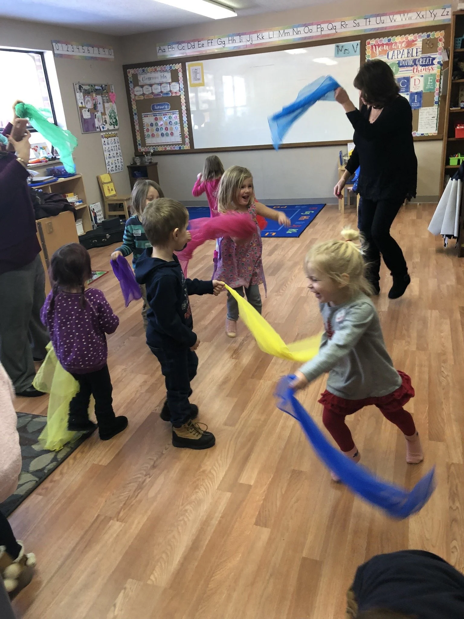 Young children are dancing and playing with colorful scarves in a classroom, with a teacher leading the activity.