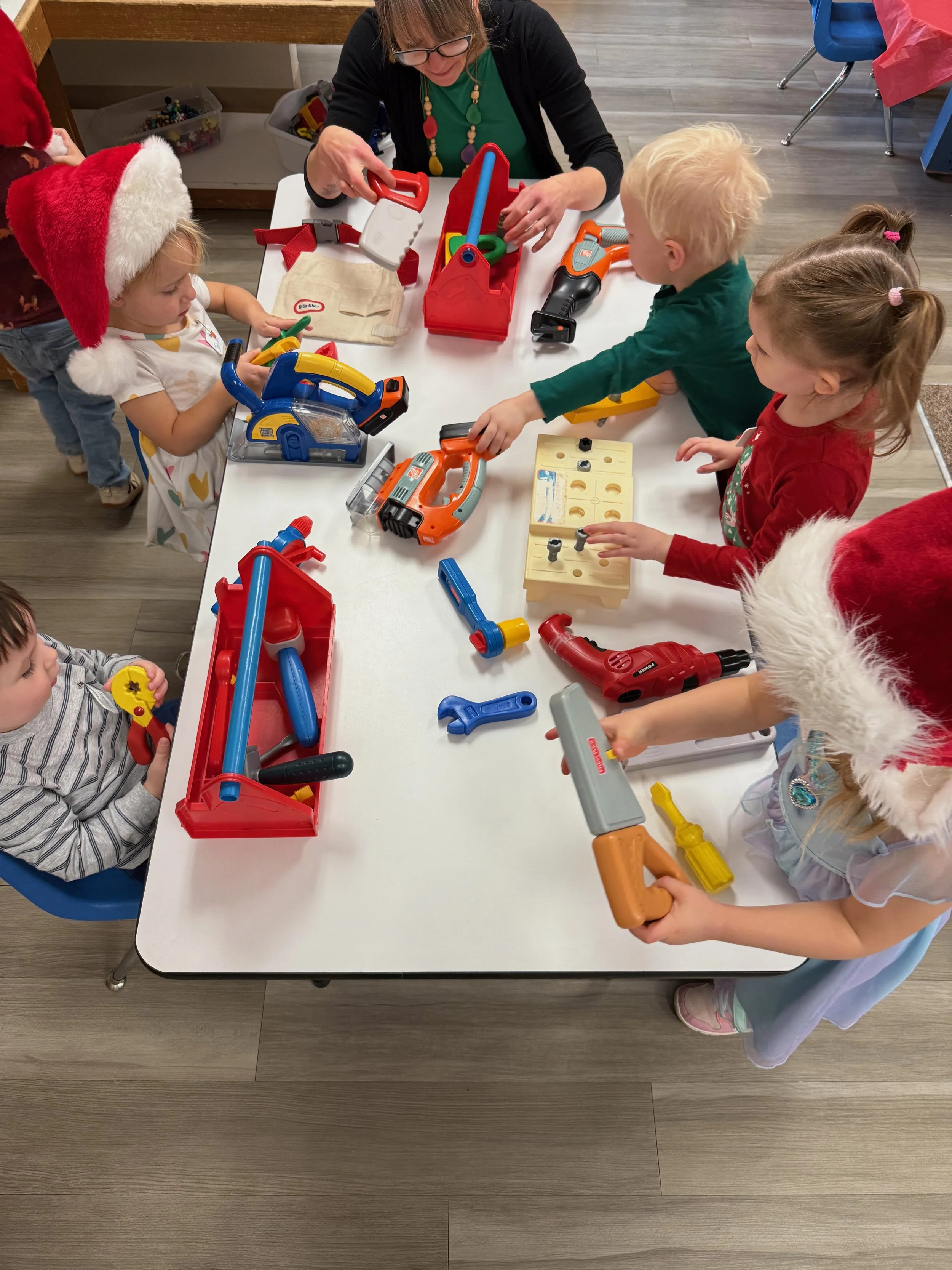 Children playing with toy tools and power tools at a table, some children are wearing Santa hats, in a classroom or playroom setting.