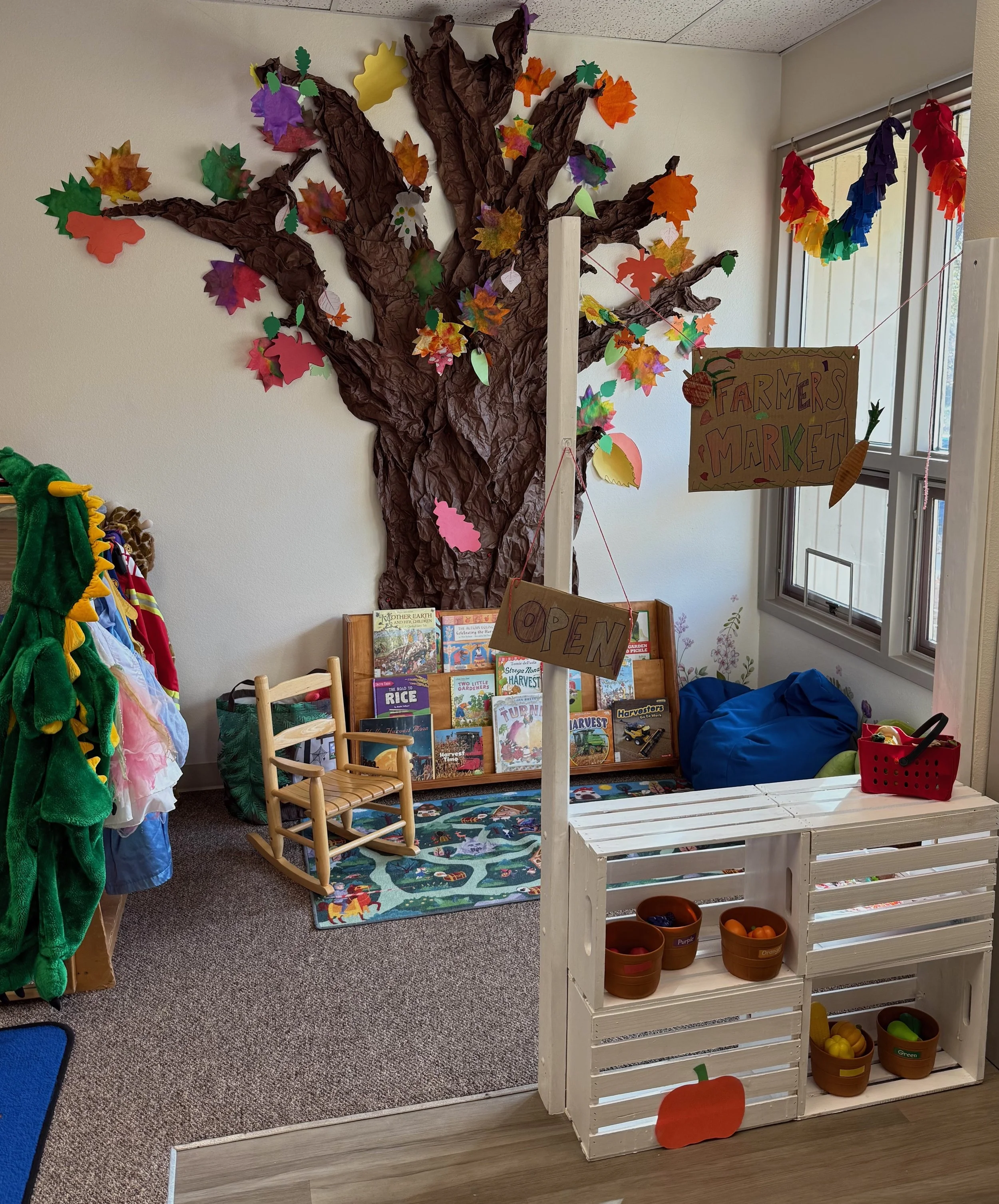 A mini farmers market setup for children with a large paper tree decoration on the wall, a small rocking chair, and shelves with toy fruits and books.