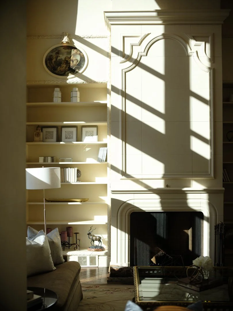 Living room with a built-in white bookshelf and fireplace, sunlight casting shadows across the space, decorative items on shelves, a beige sofa with pillows, and a glass-topped coffee table.